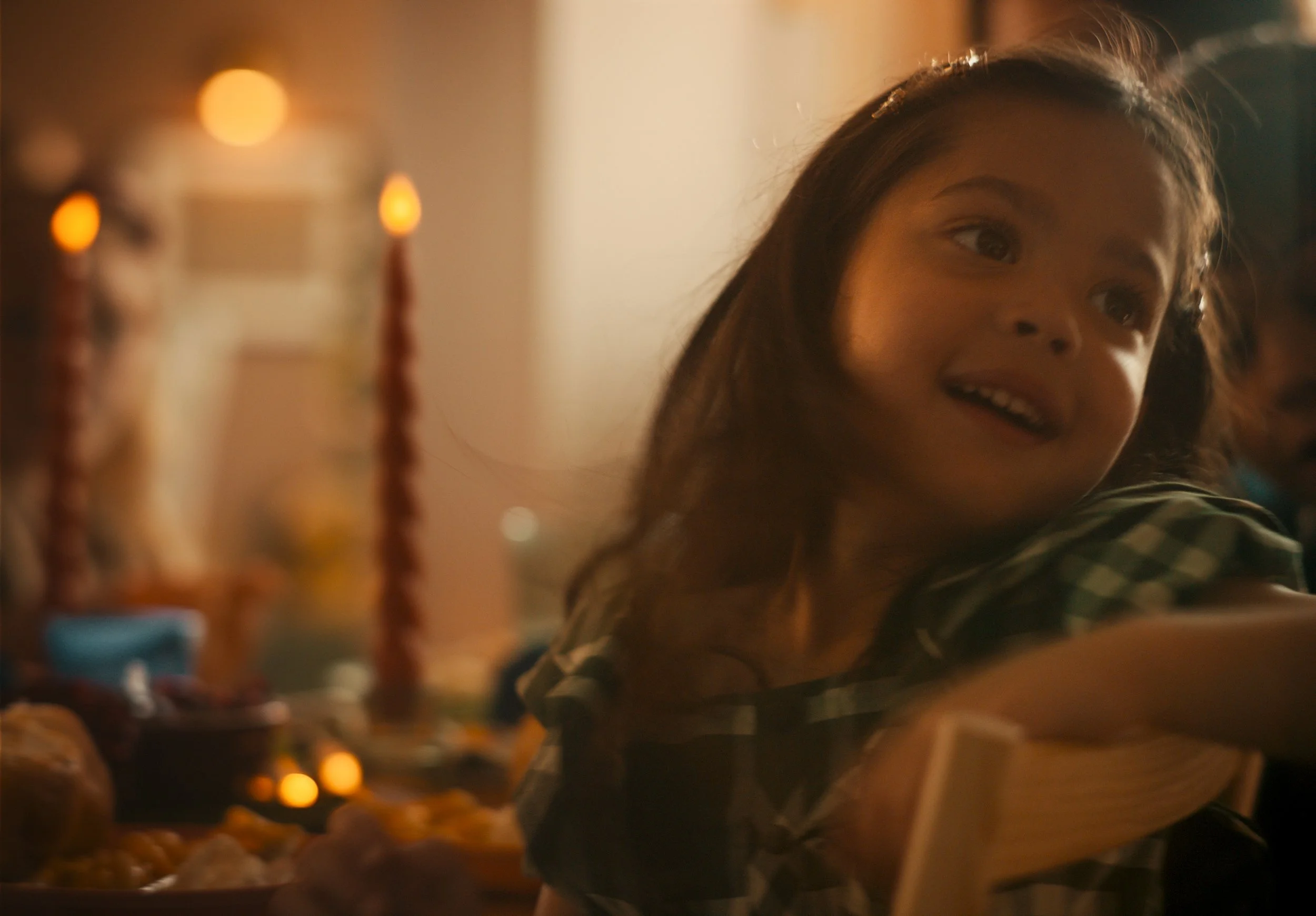 A young girl with long dark hair at a birthday celebration, smiling and looking to the side, with birthday candles and cake visible in the background.