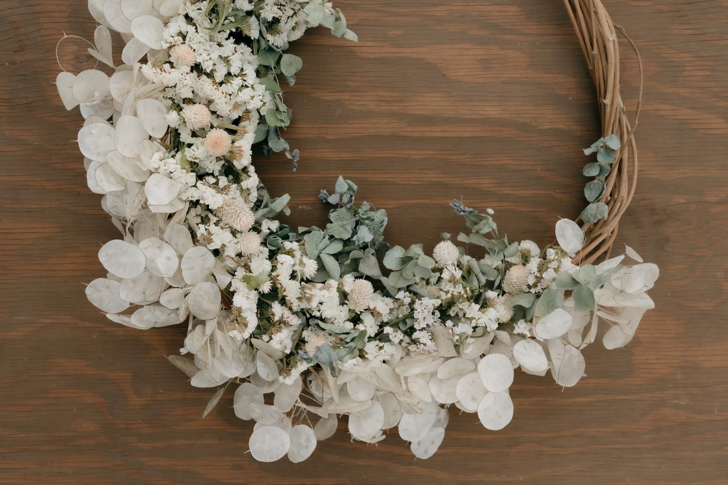 A partially completed wreath made of white dried flowers and eucalyptus leaves, resting on a wooden surface.
