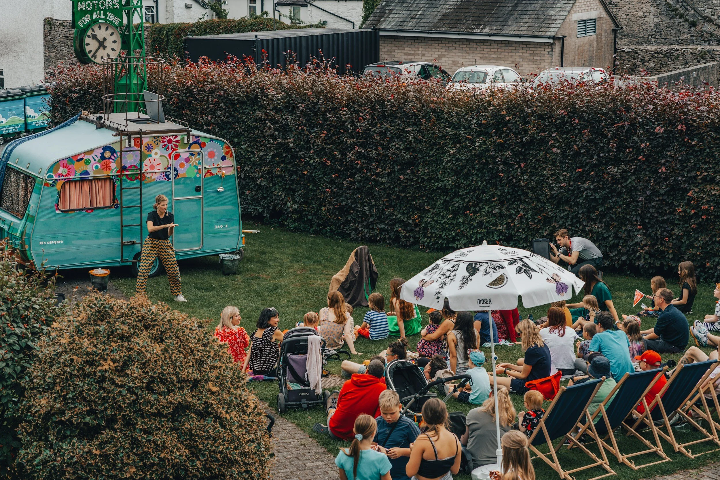 Outdoor event in a garden with a vintage turquoise caravan, decorated with flowers, serving as a backdrop. A crowd of adults and children sit on the grass, some in deck chairs, and gather under an umbrella, watching a performance or activity. The setting has a relaxed atmosphere with hedges and a parking area in the background.