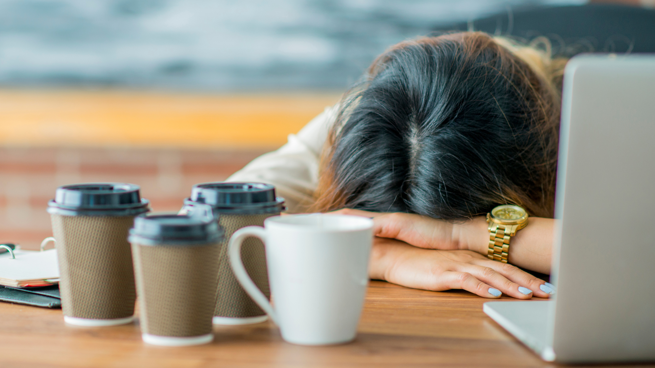 woman collapsed in exhaustion with head on desk by laptop with several cups of coffee on her desk