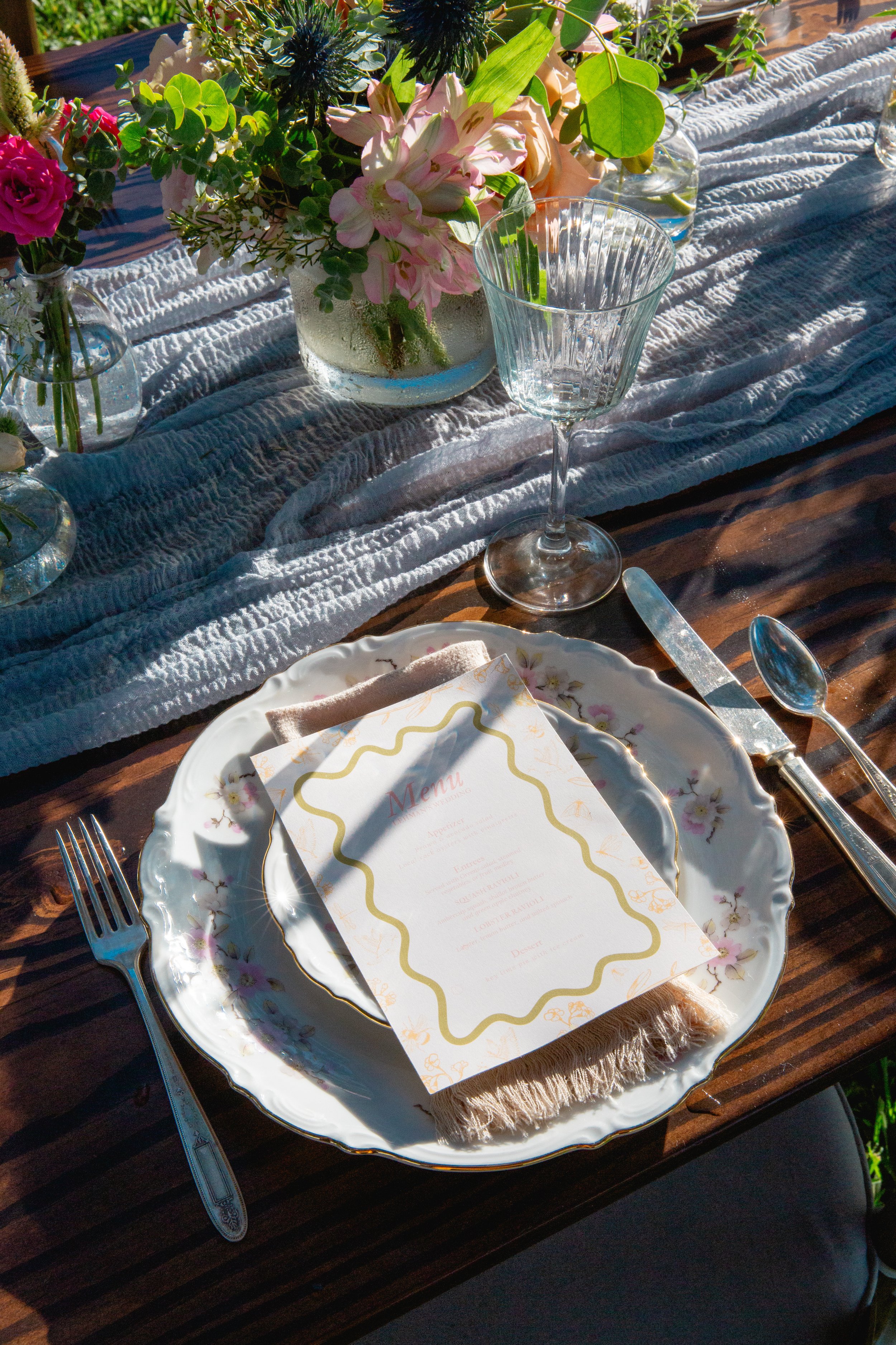A table setting with a floral centerpiece, a vintage floral-patterned plate with a folded placemat, a menu card, silverware, and an empty wine glass, arranged on a dark wooden table with a light cloth runner.