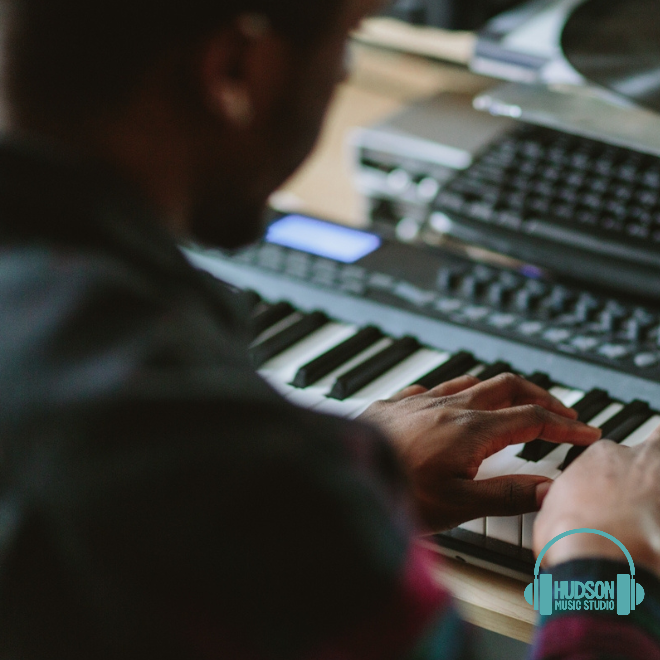 Person playing keyboard at a music studio with laptops and music equipment nearby, logo of Hudson Music Studio in bottom right corner.