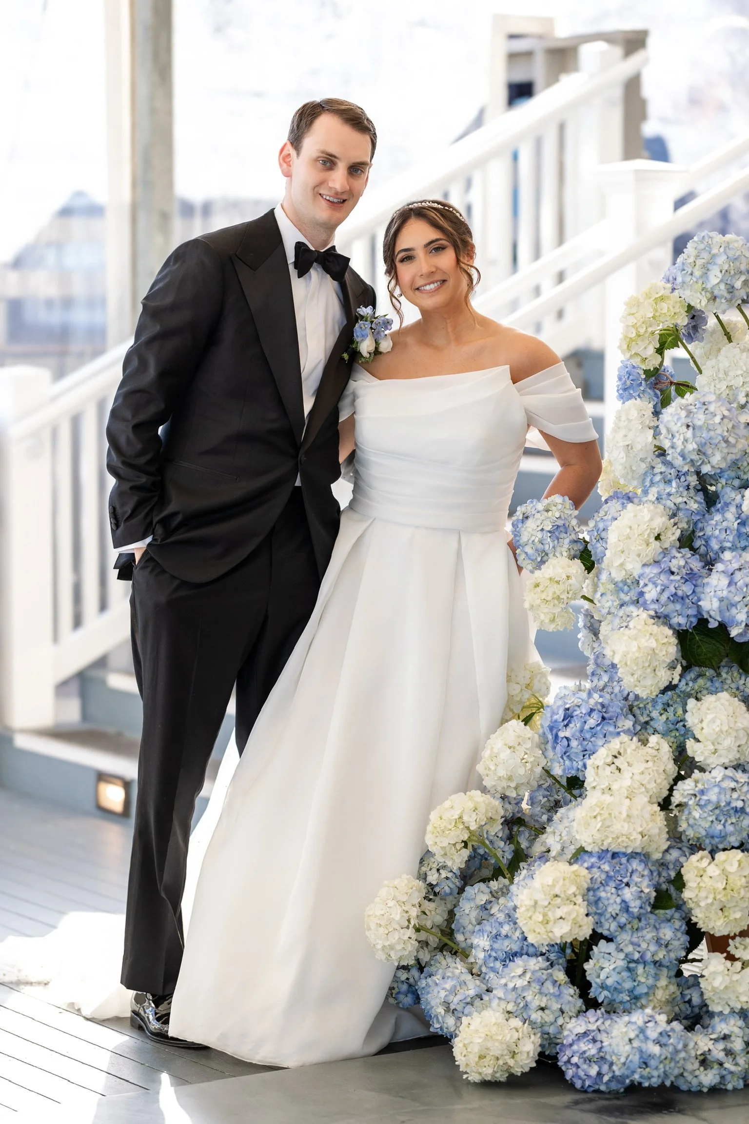 A happy wedding couple standing next to a large arrangement of blue and white hydrangeas, inside a bright indoor venue.