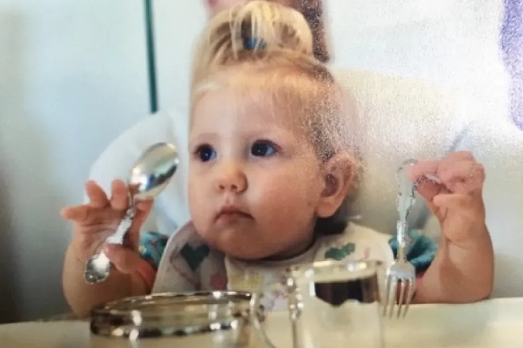 Baby Liv eating at a high chair with silverware, rocking a top-knot and bib.
