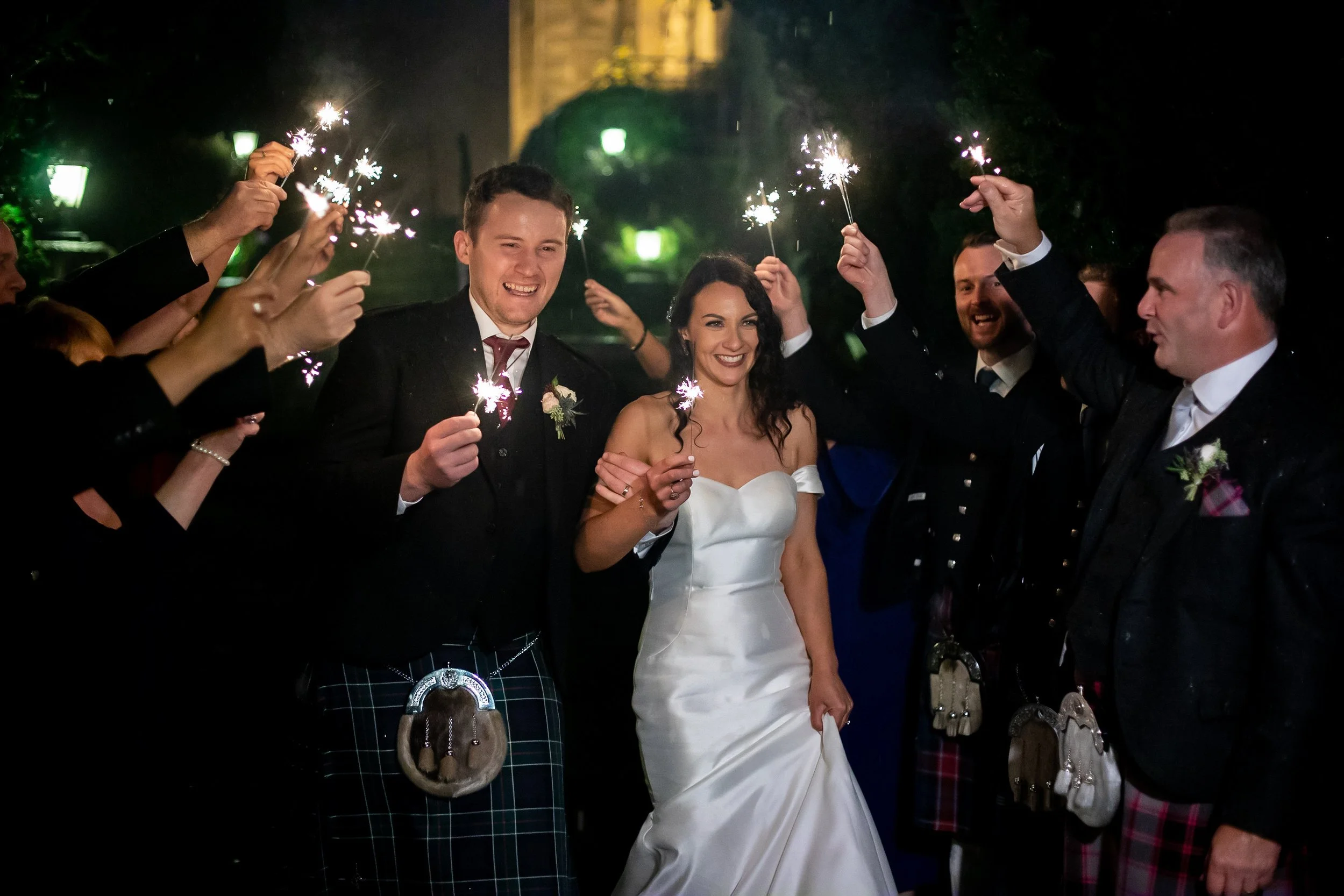 Wedding couple holding sparklers surrounded by guests in evening attire, smiling and celebrating.
