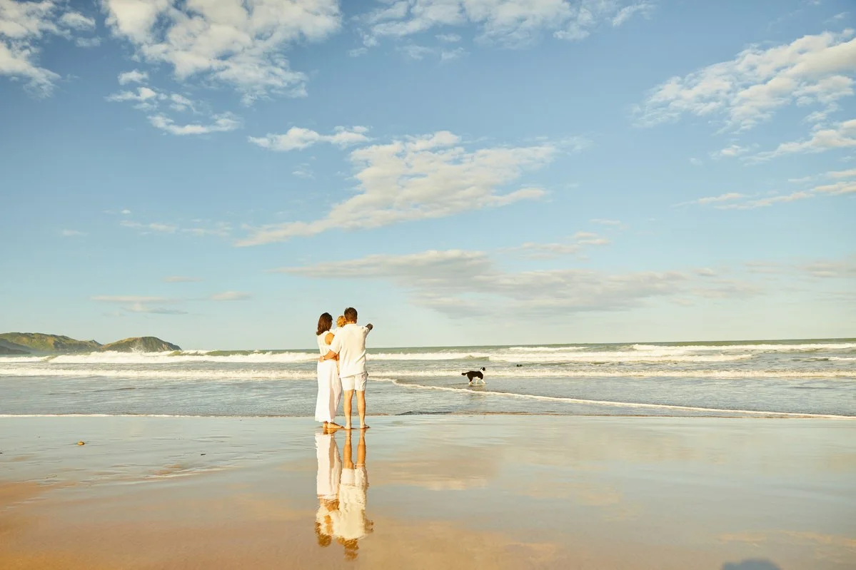 Family standing on a beach with the ocean and distant hills in the background, reflecting in the wet sand.