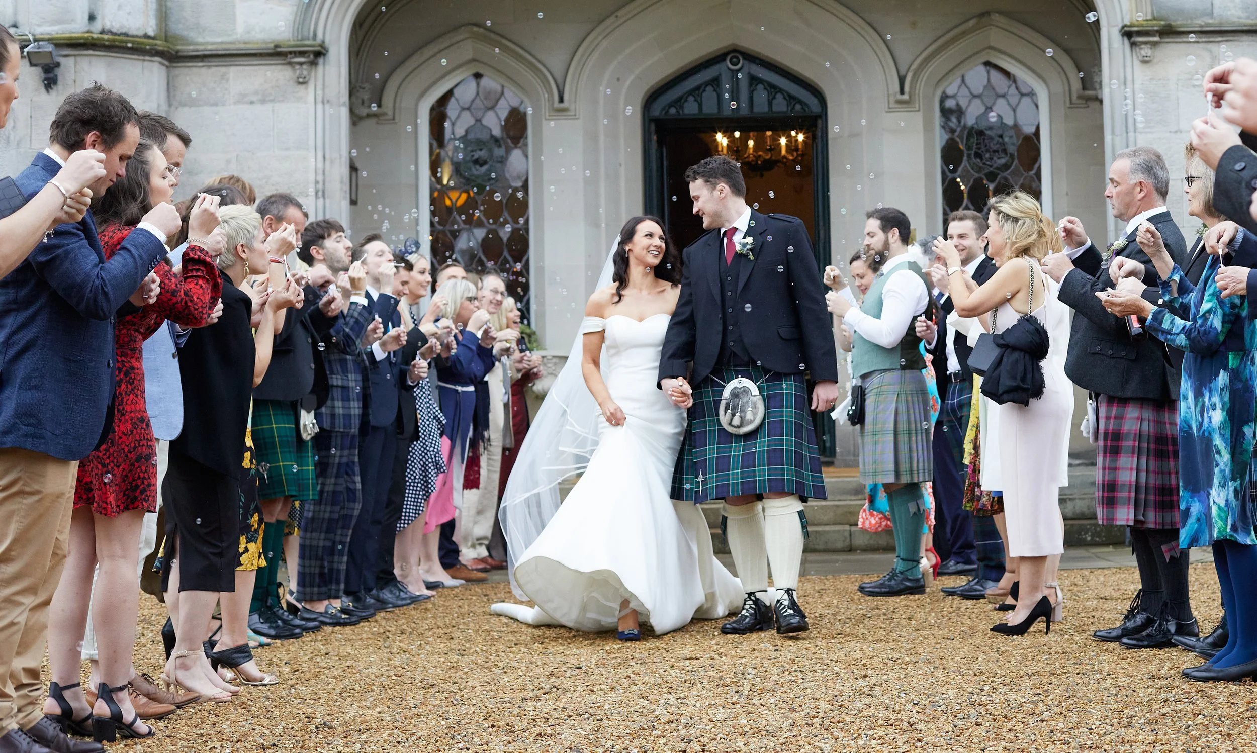 Bride and groom walking through a crowd at a wedding, with guests blowing bubbles, near a stone building with arched doorways.