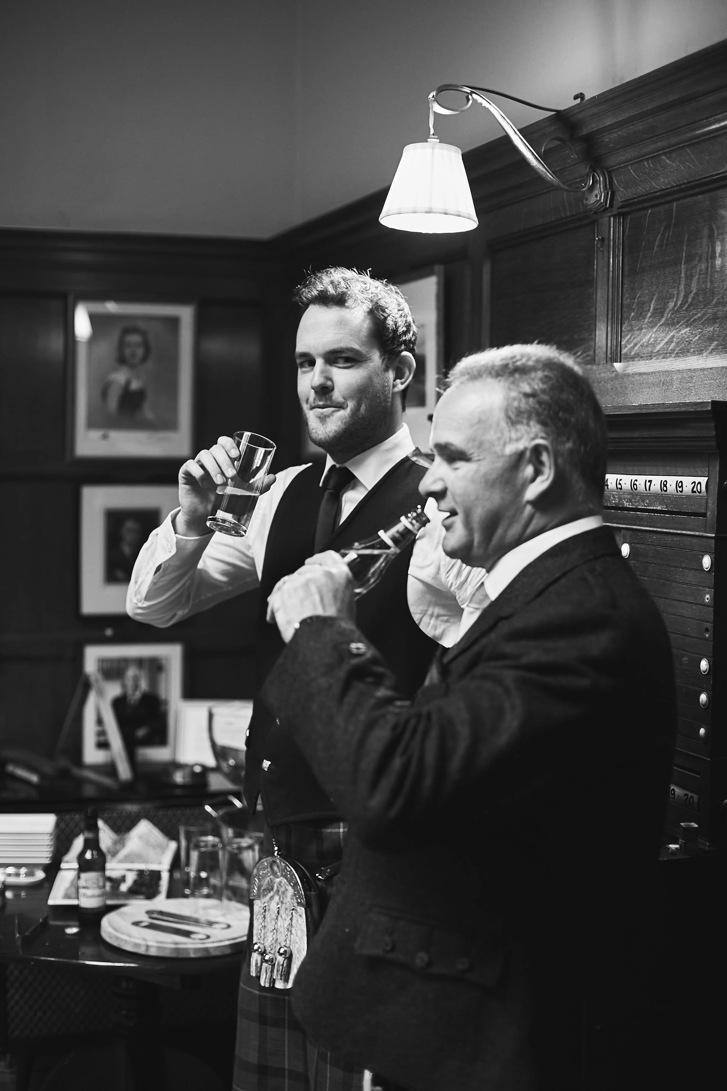 Two men in formal attire enjoying drinks in a room with framed photos and a lamp.
