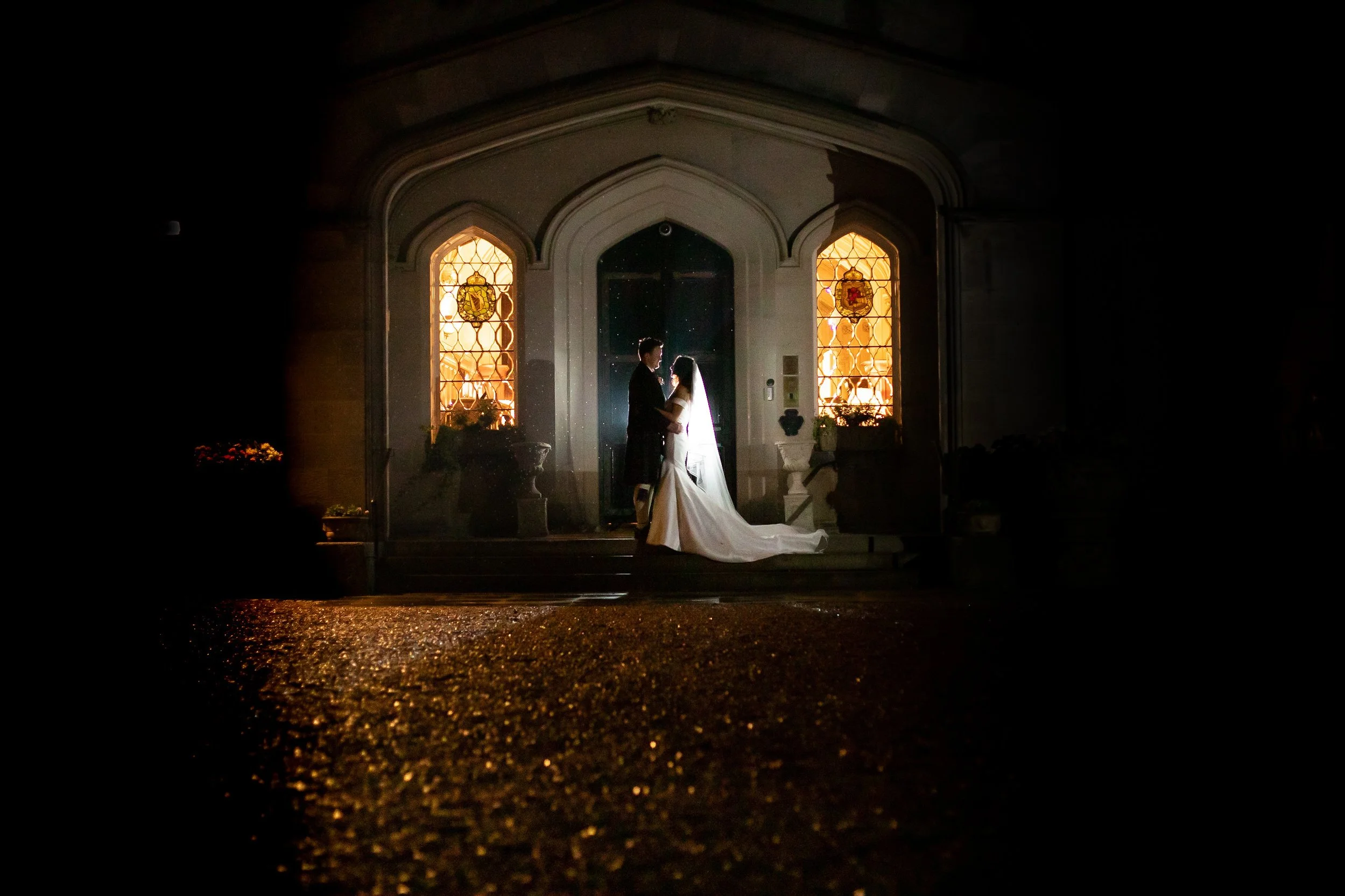 Bride and groom at night in front of building with stained glass windows, illuminated by warm light.