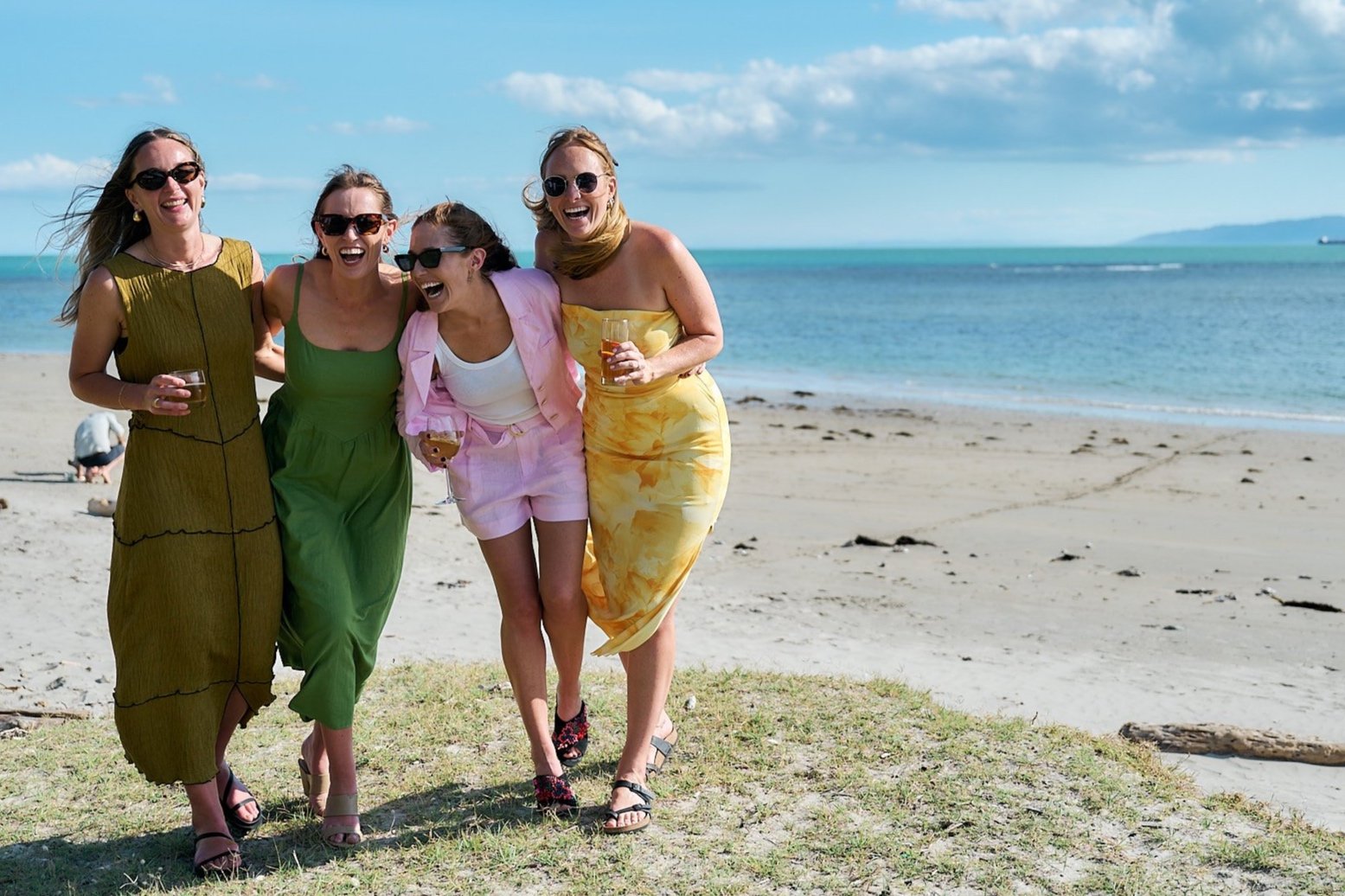 Four women laughing and walking on a beach with the ocean in the background.