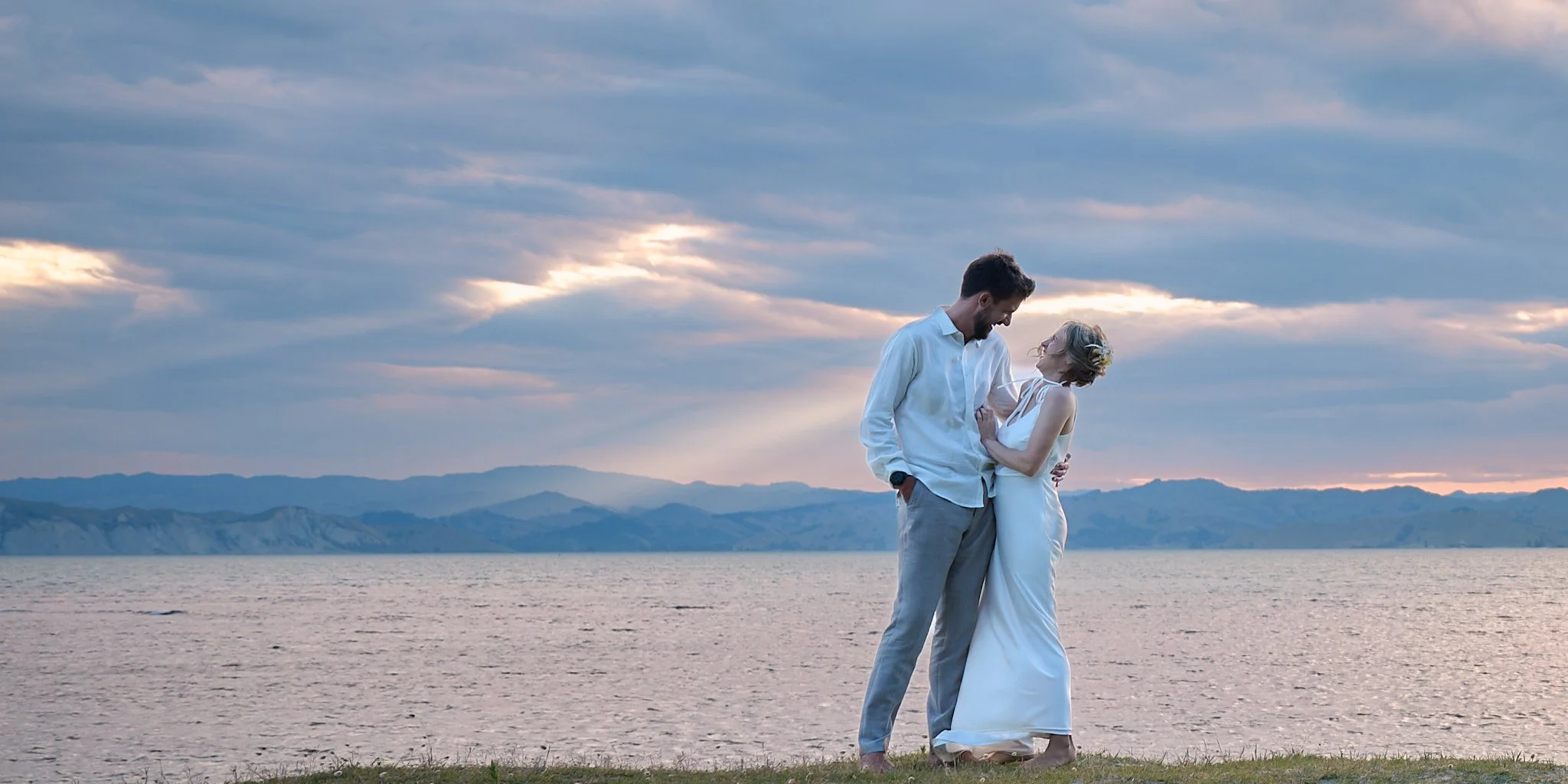 A couple embracing on a beach at sunset, with mountains and sea in the background.