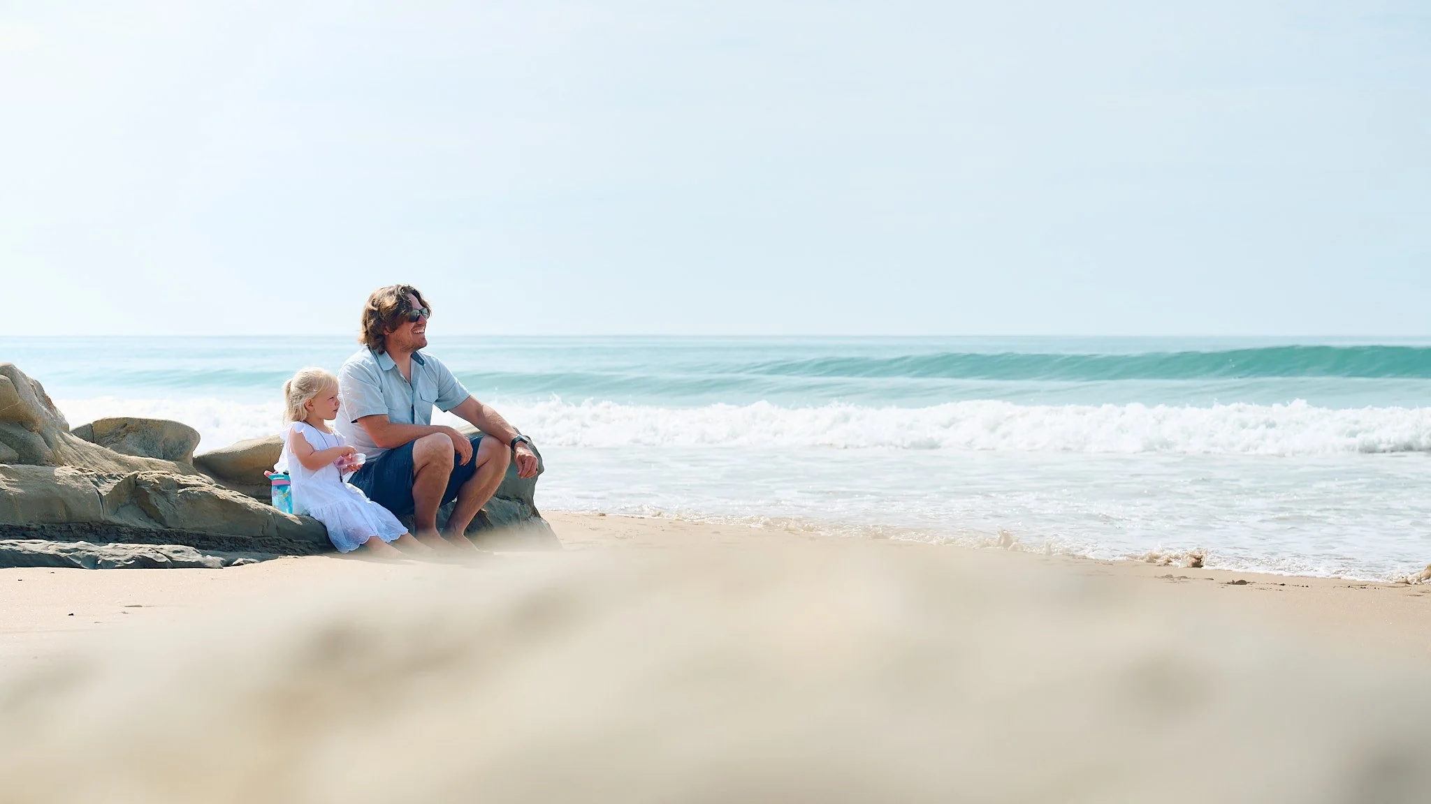 Man and child sitting on a beach near rocks, looking at the ocean waves.
