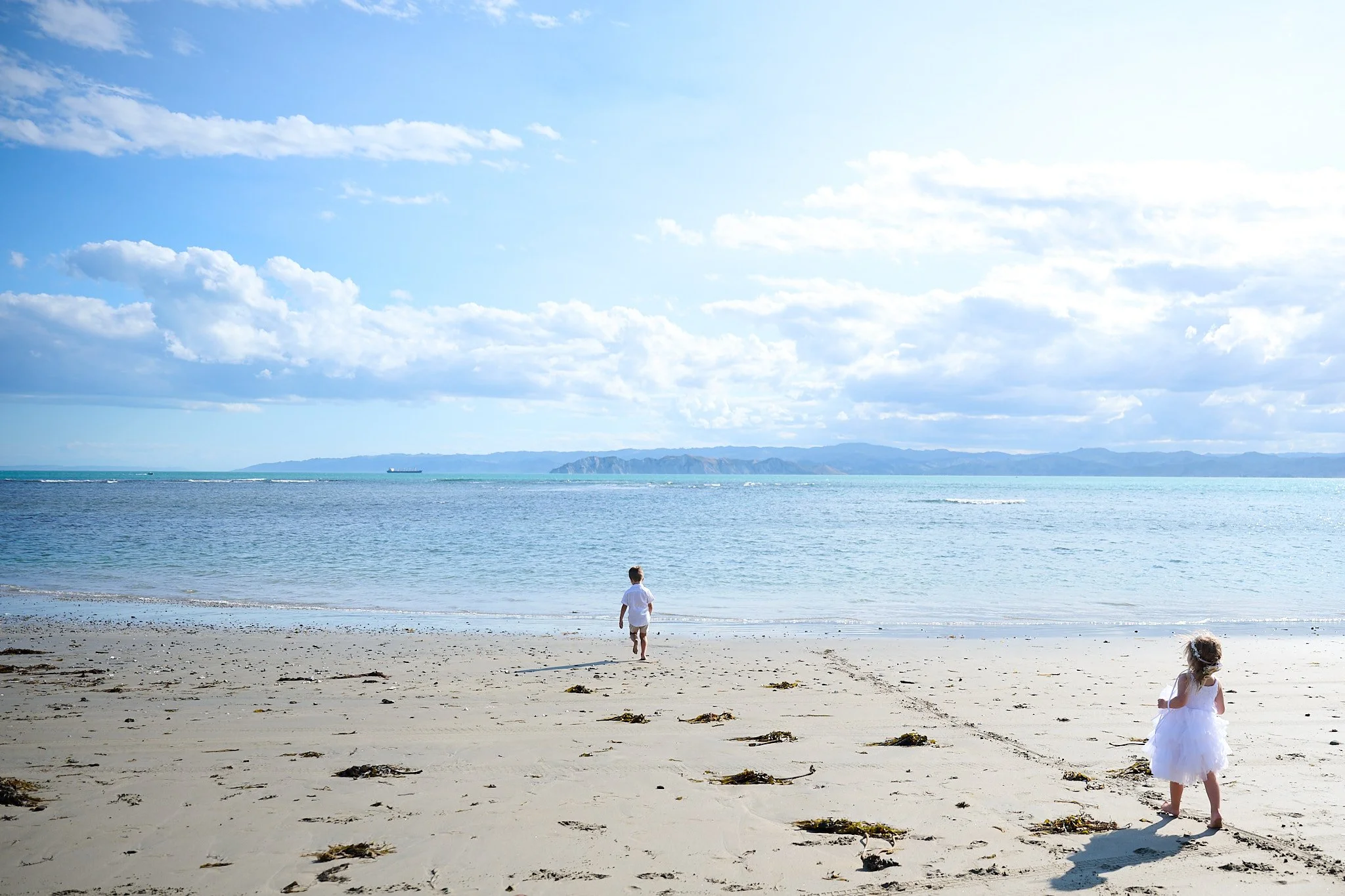 Two children walking on a sandy beach towards the ocean, with a clear blue sky and distant mountains in the background.