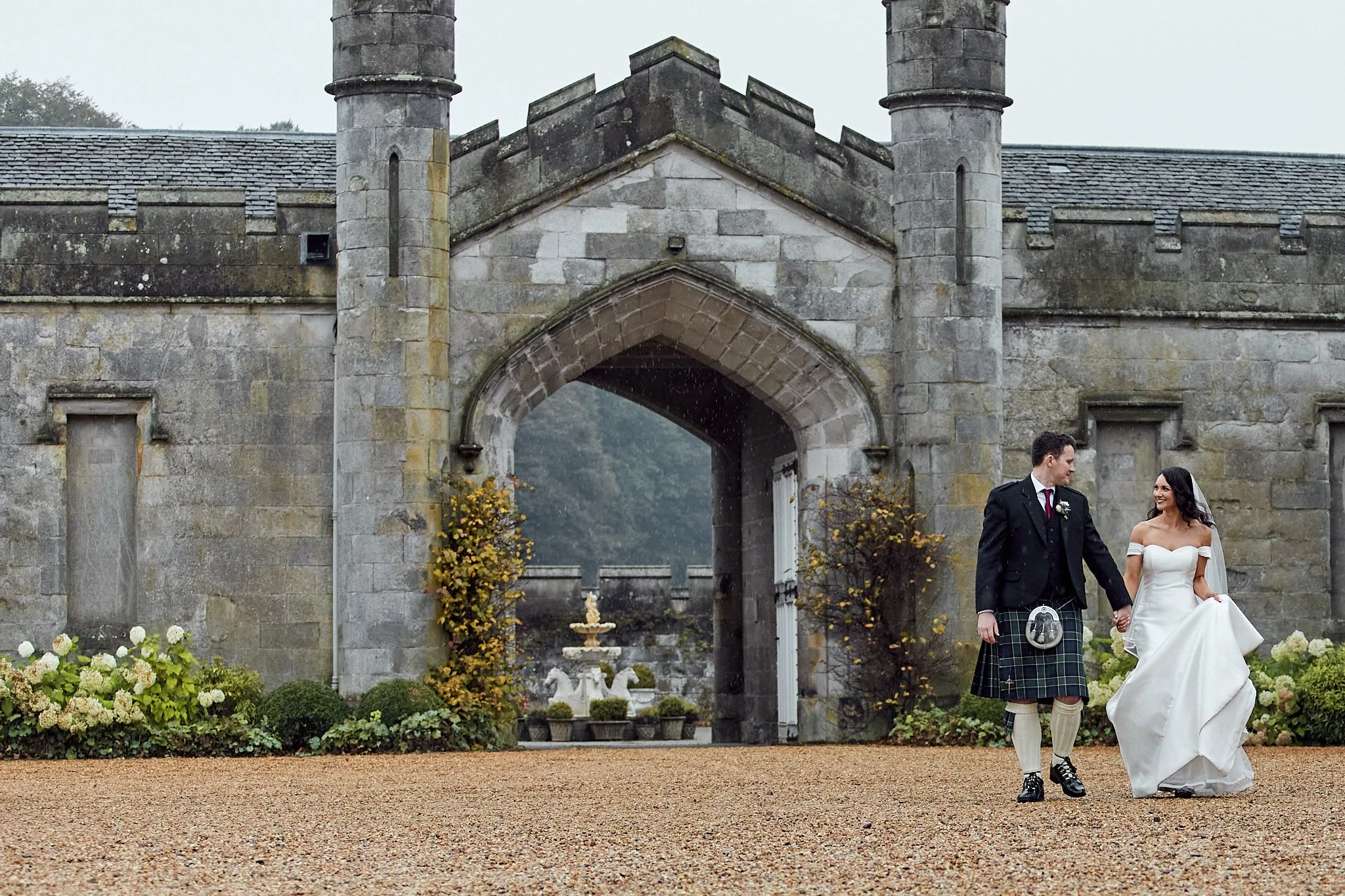 Bride and groom walking hand in hand in front of a historical stone building with arches and a garden.