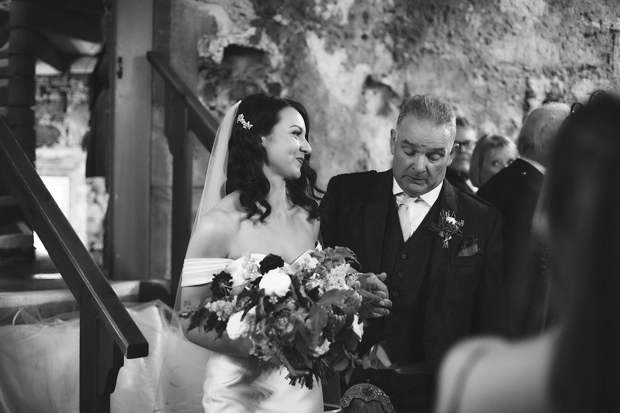 Bride holding bouquet, escorted by a man in a formal setting.