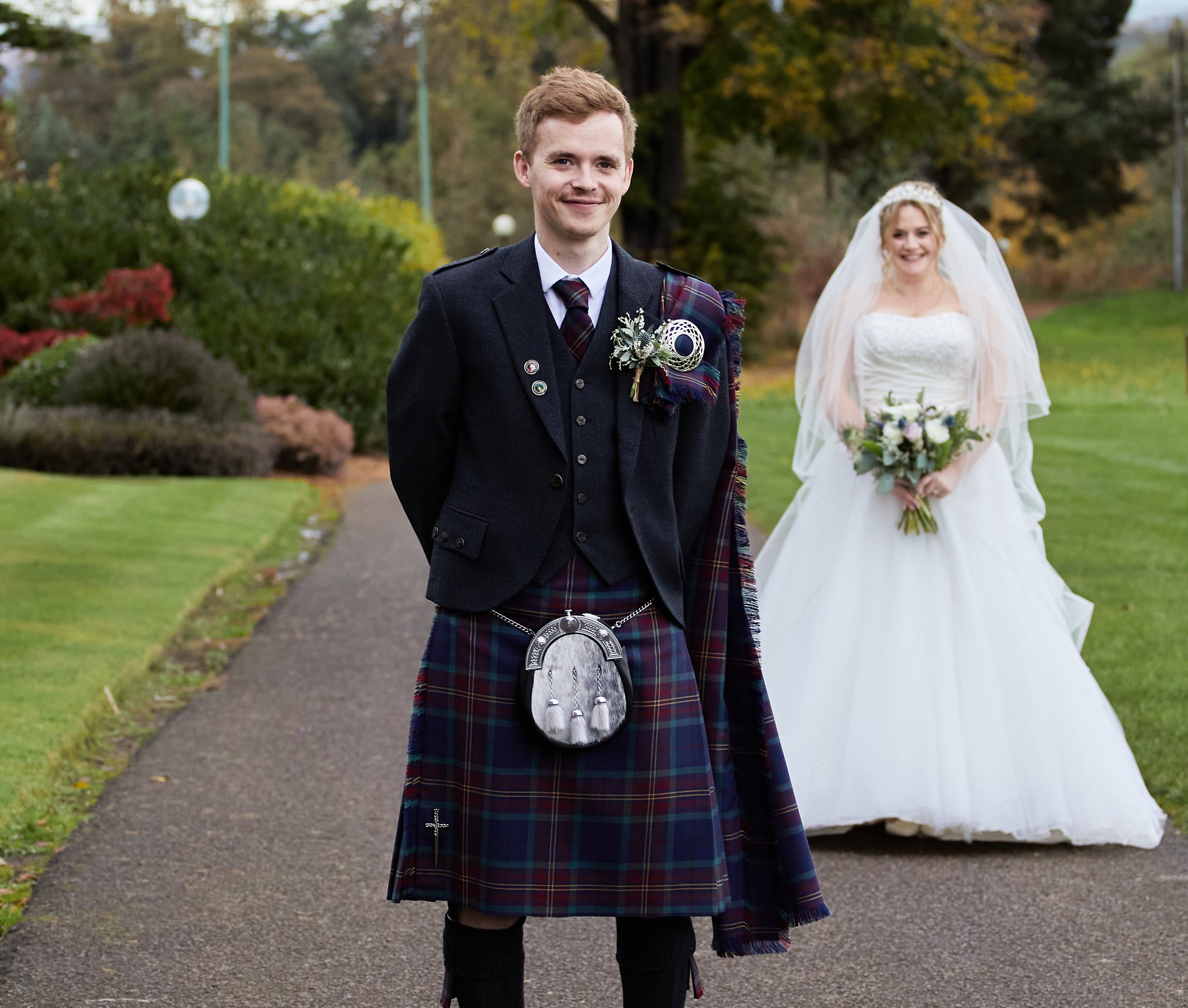 A groom in a traditional Scottish kilt, with a bride in a wedding dress holding a bouquet, standing outdoors on a path.