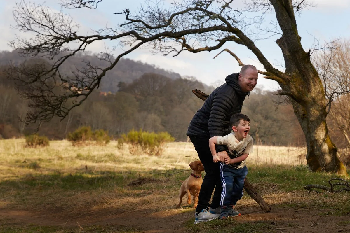 A man and a child playing with a dog in a scenic outdoor setting, featuring a tree and hills in the background.