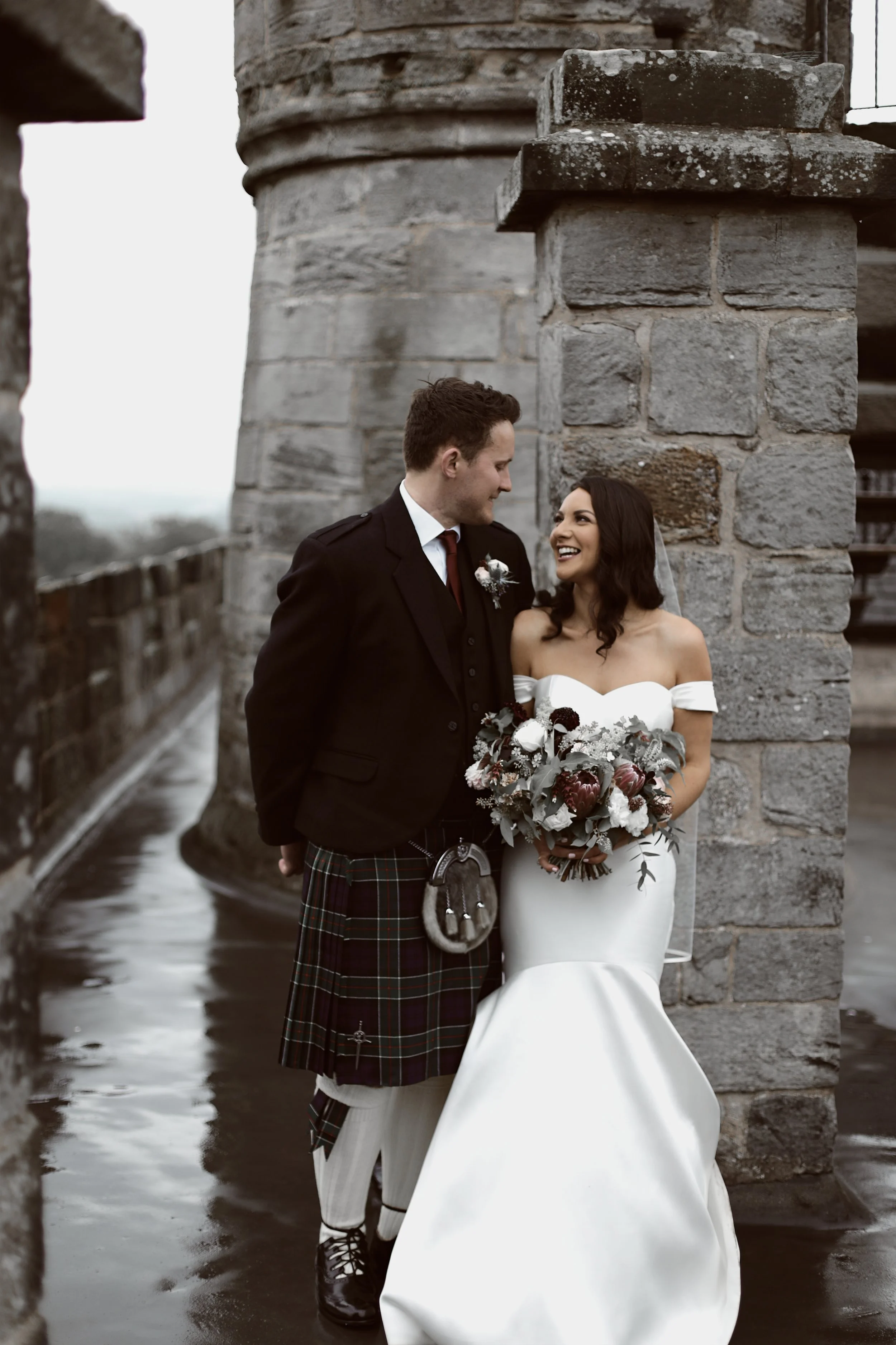 Bride and groom in traditional attire pose near stone wall, groom in kilt, and bride in white dress holding bouquet.