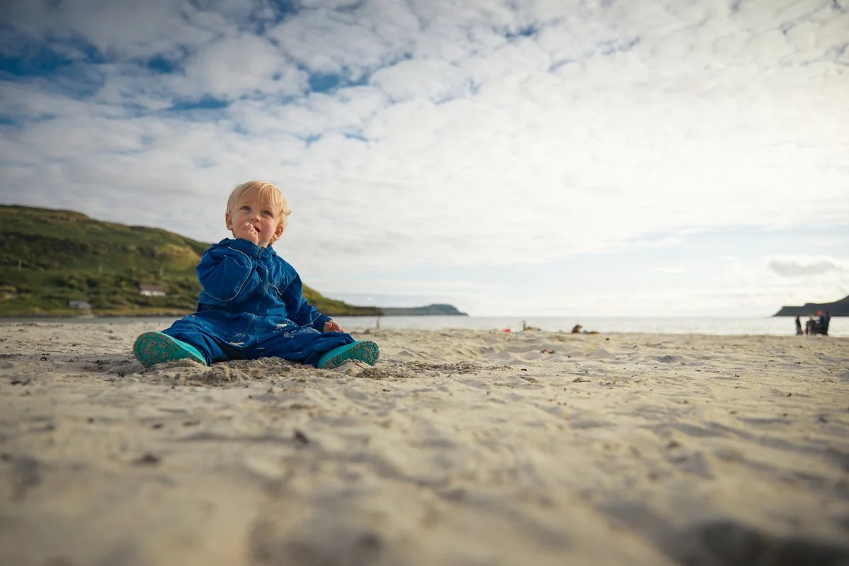 Toddler in blue snowsuit sitting on a sandy beach with distant hills and cloudy sky.