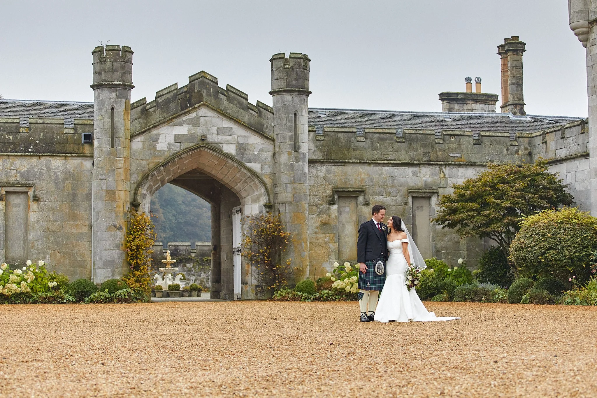 Bride and groom standing in front of a stone castle with a fountain in the background.