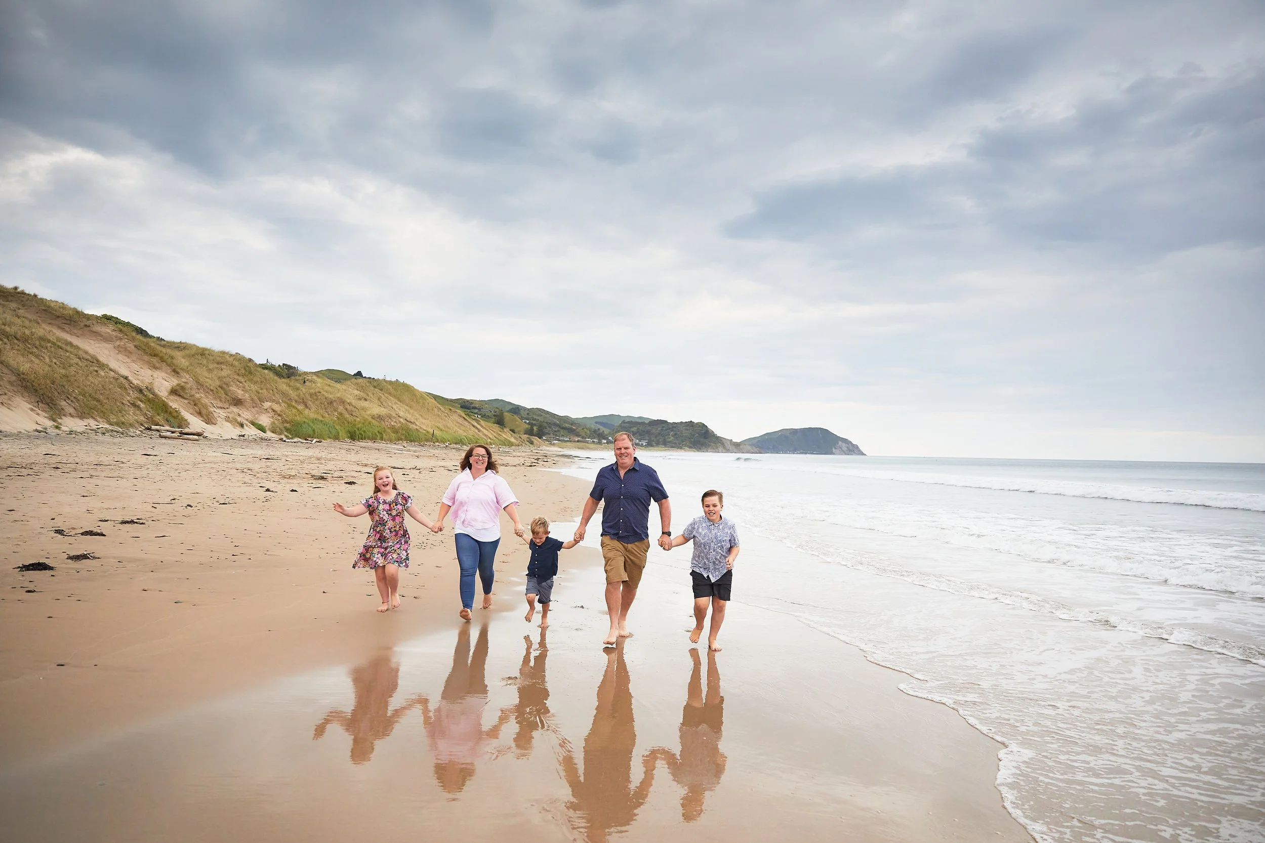Family walking on a beach holding hands, with ocean and hills in the background.
