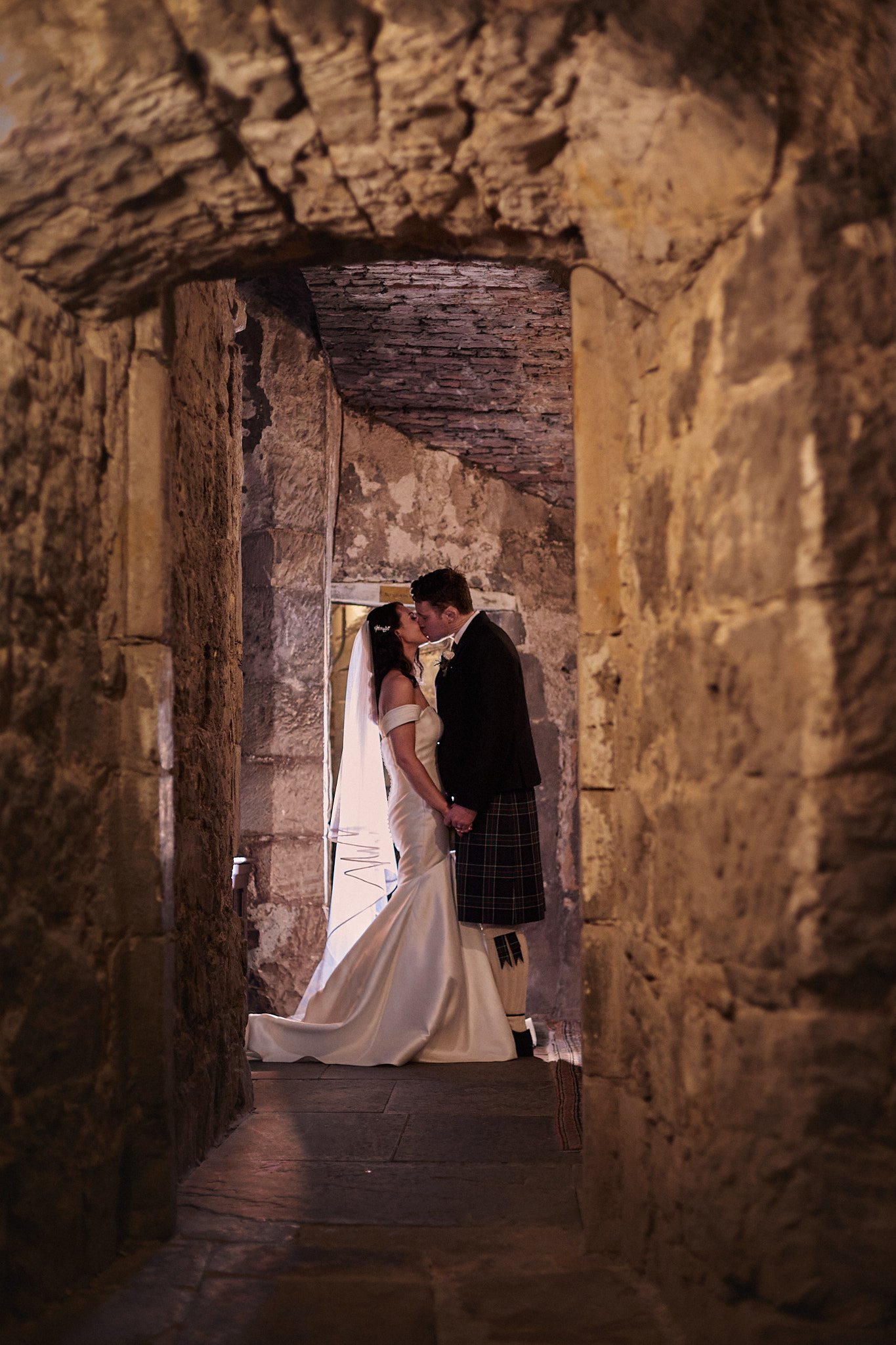 A groom in a kilt kissing a bride in a wedding dress in a stone corridor.