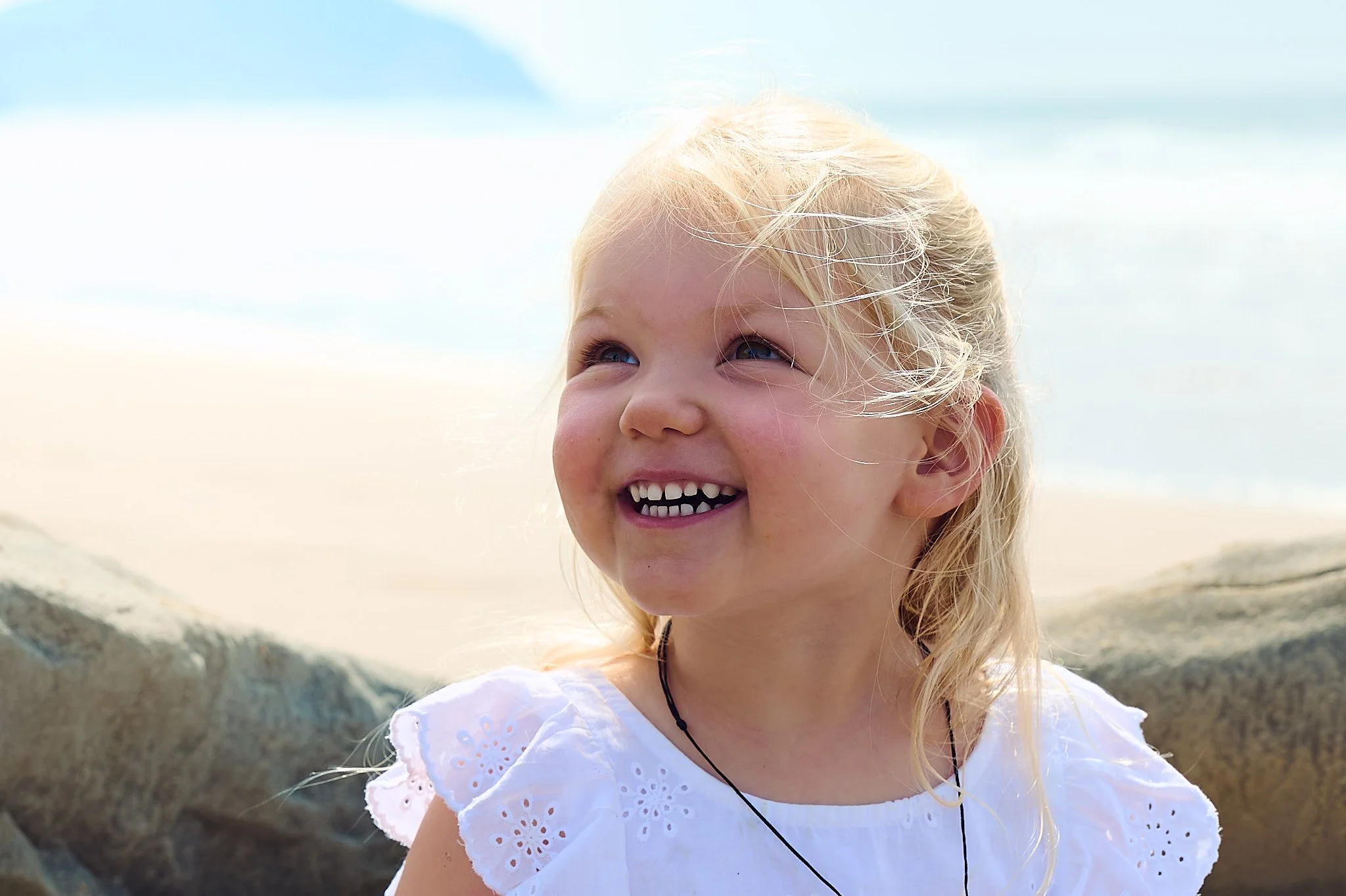 Smiling child with blonde hair at the beach, wearing a white dress.