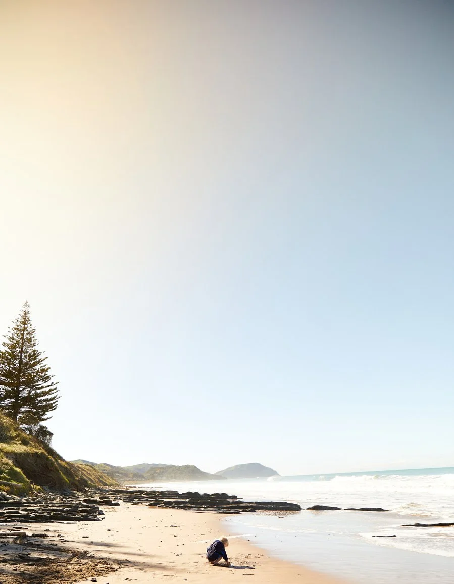 Child playing on a sandy beach near the ocean with rocks and a tree in the background under a clear sky.