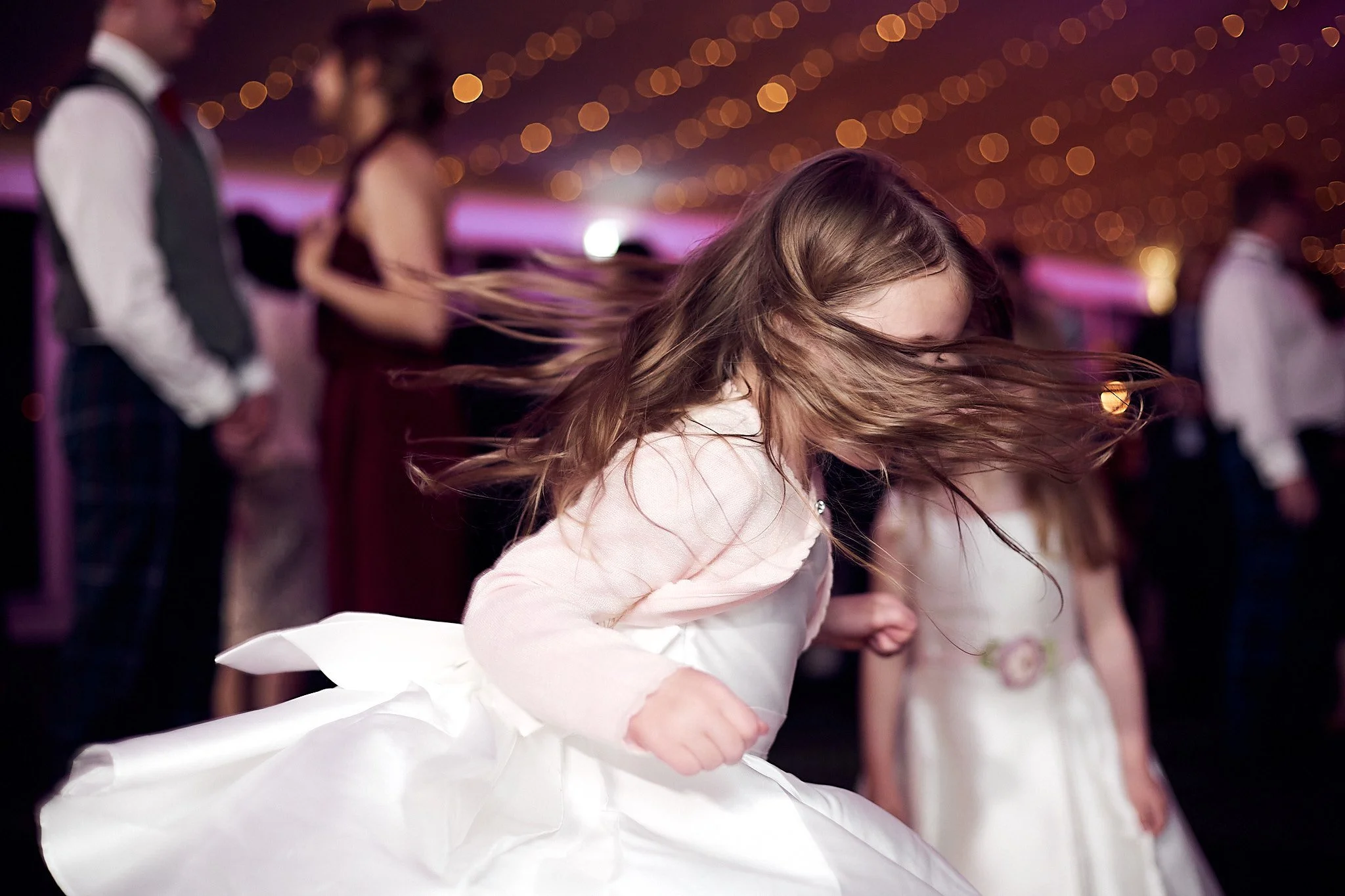 Young girl in white dress dancing at a festive event with blurry background and string lights.