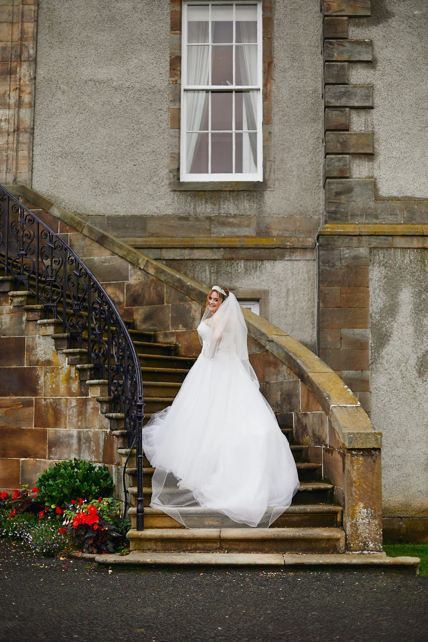 Bride in a white wedding gown on stone stairs next to a historic building with a window, surrounded by red flowers and greenery.