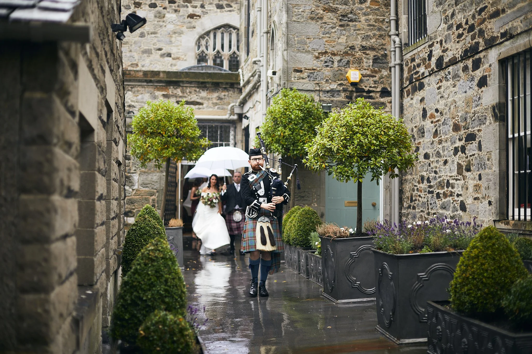 A bagpiper in traditional Scottish attire plays in front of a stone building, followed by a bride and groom under an umbrella, walking through a courtyard with greenery.