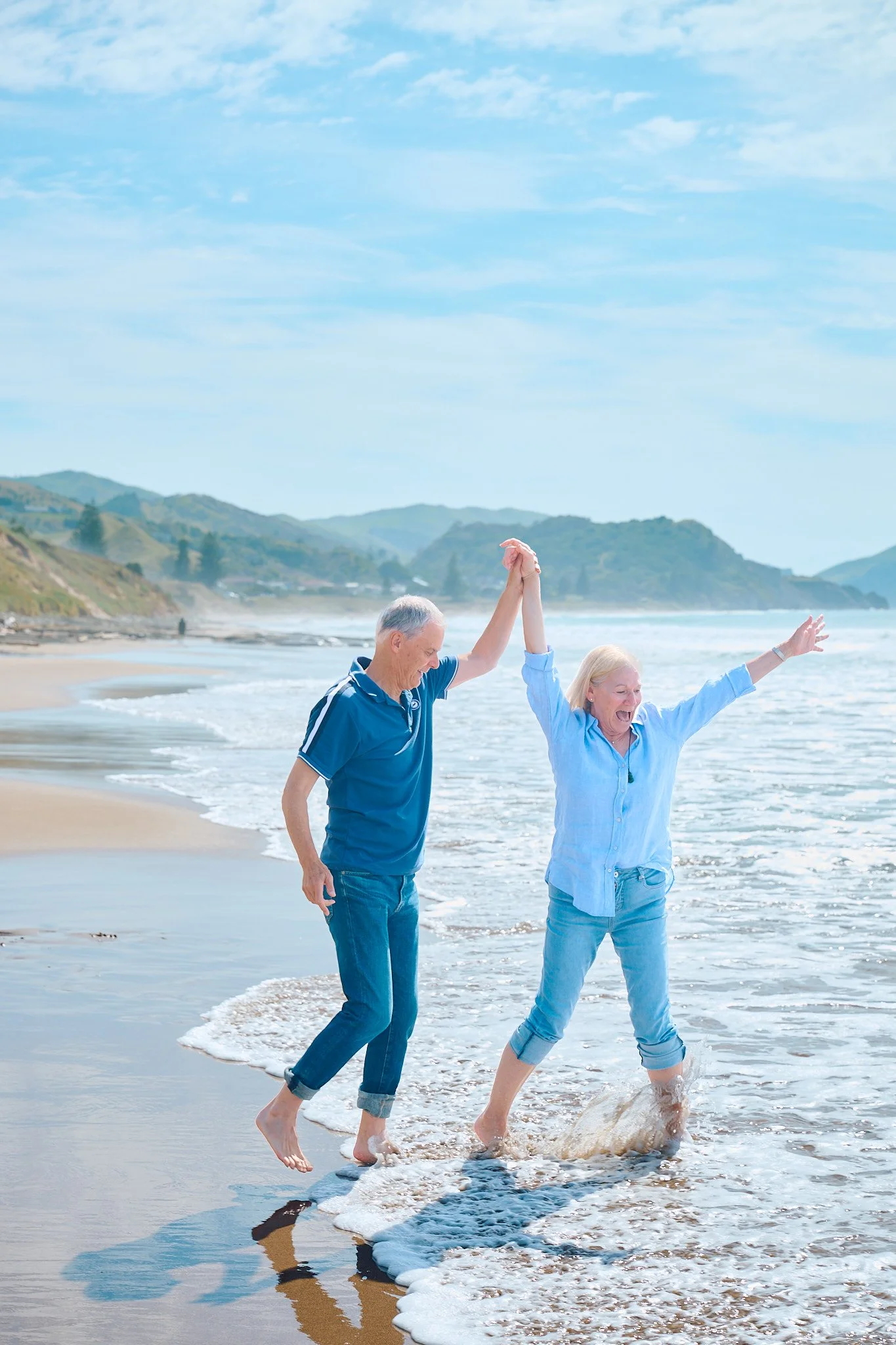 Senior couple joyfully walking along a sunny beach, holding hands, with waves splashing at their feet and hills in the background.