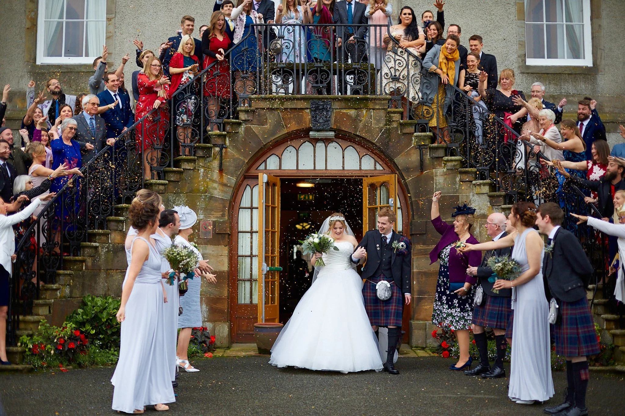 Newlyweds exiting a building under confetti, surrounded by cheering guests, with the groom in a kilt and the bride in a white gown.