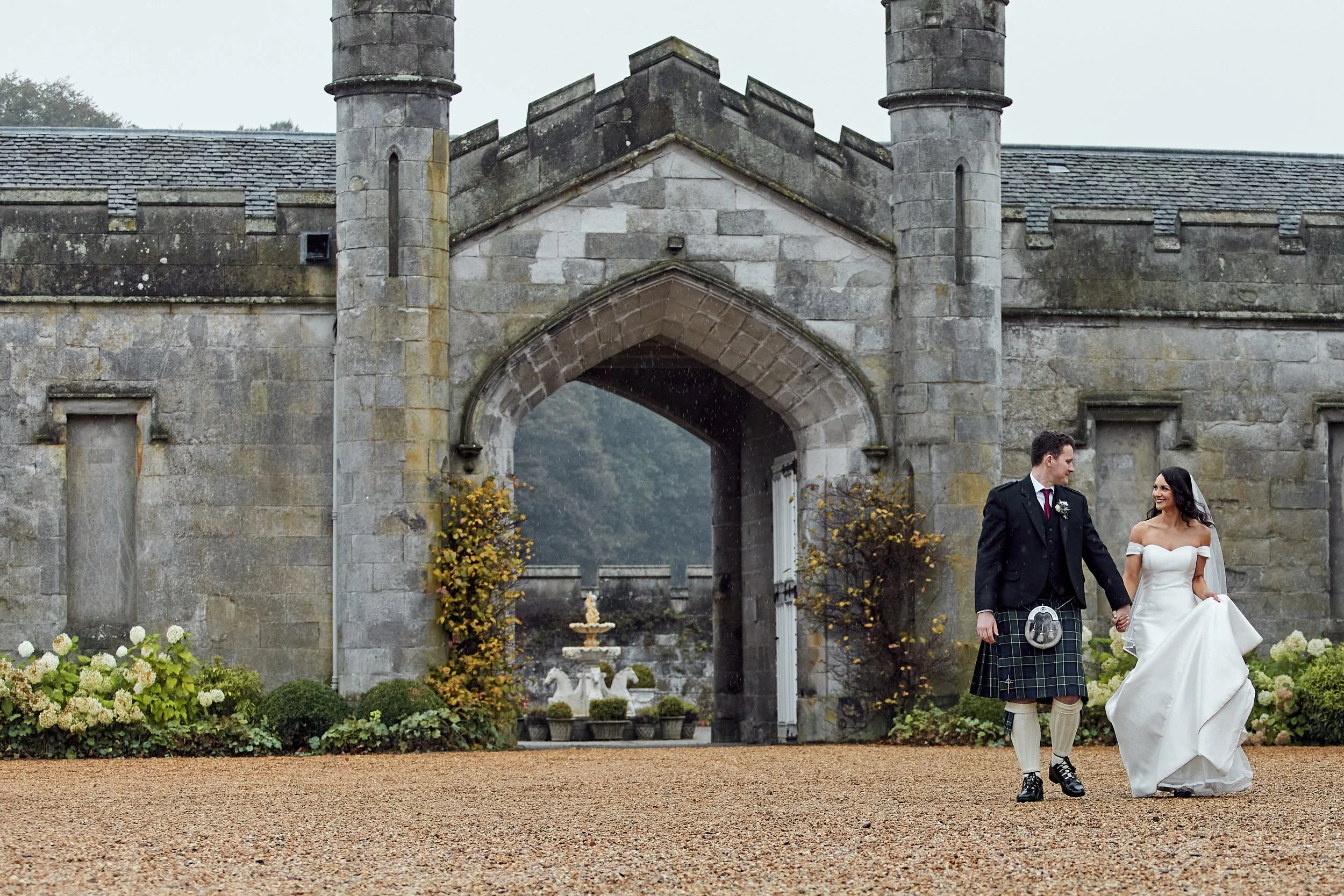 Bride and groom holding hands, walking in front of a historic stone building with an arched entrance and two pillars, surrounded by plants.