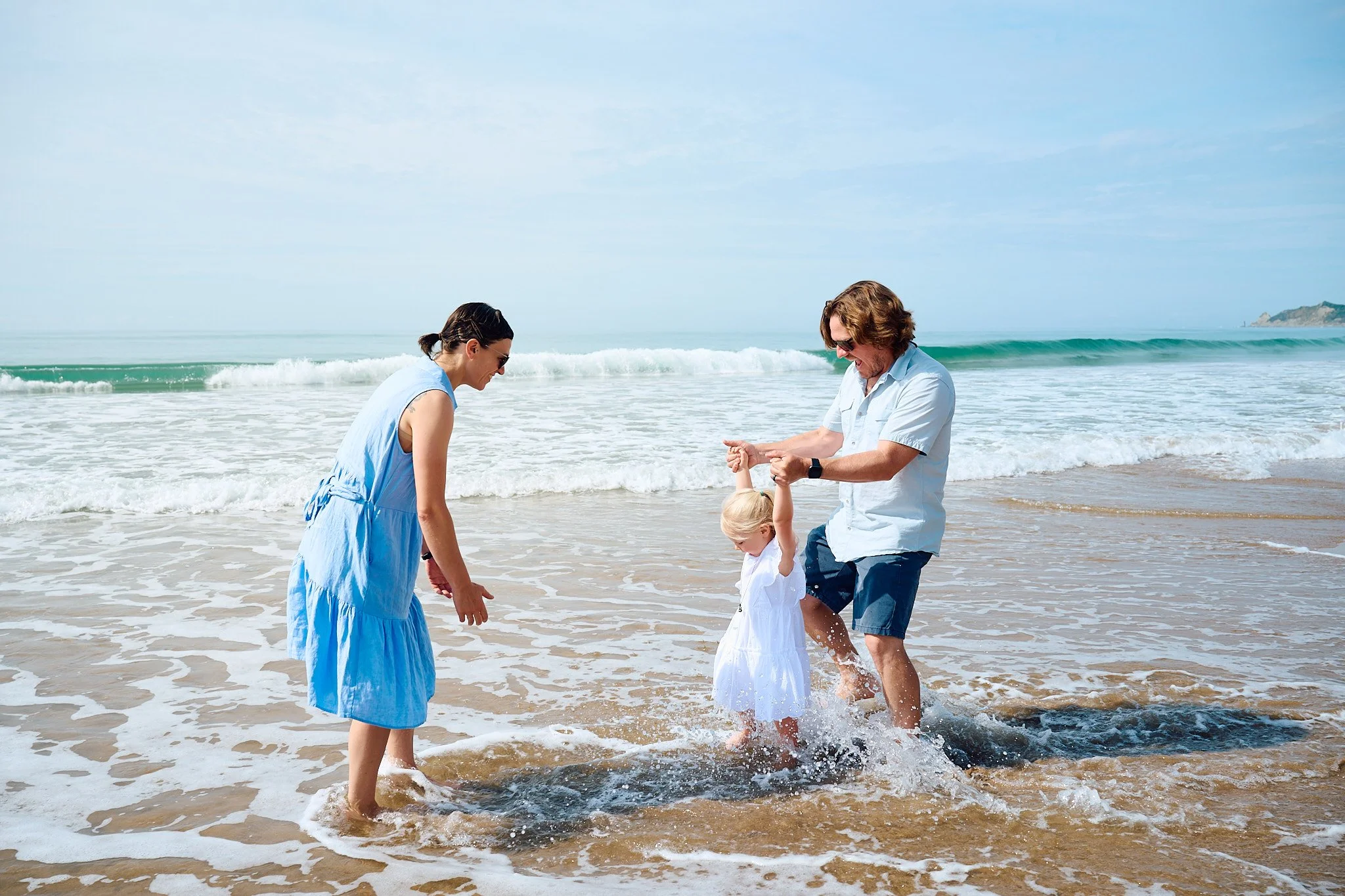 Family of three playing in ocean waves on a sandy beach.