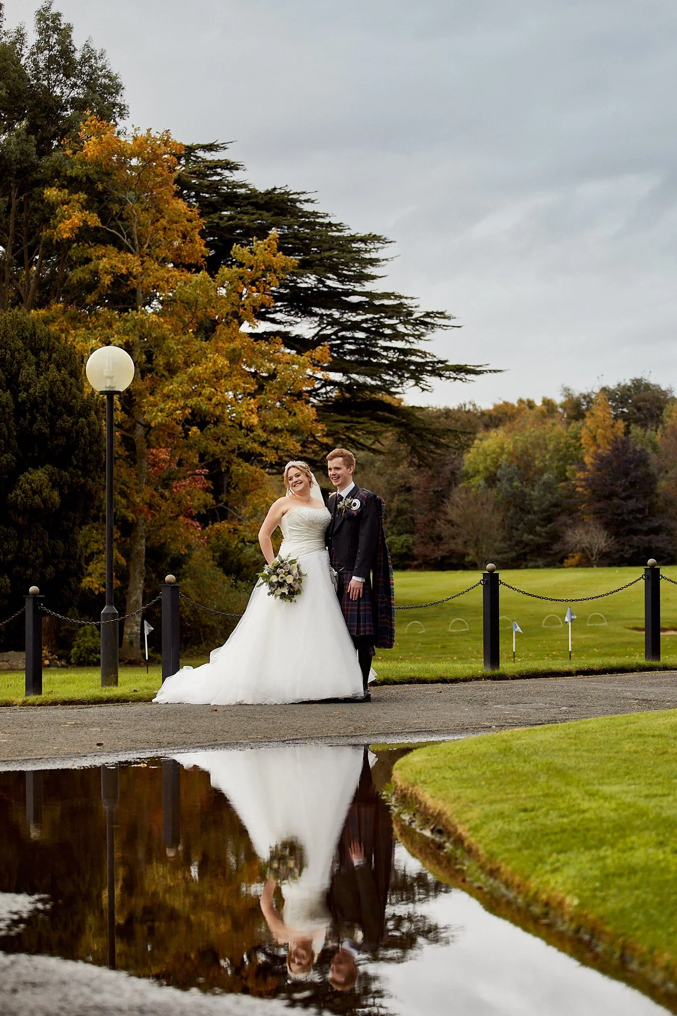Bride and groom standing outdoors near a reflection in a puddle, surrounded by autumn foliage.