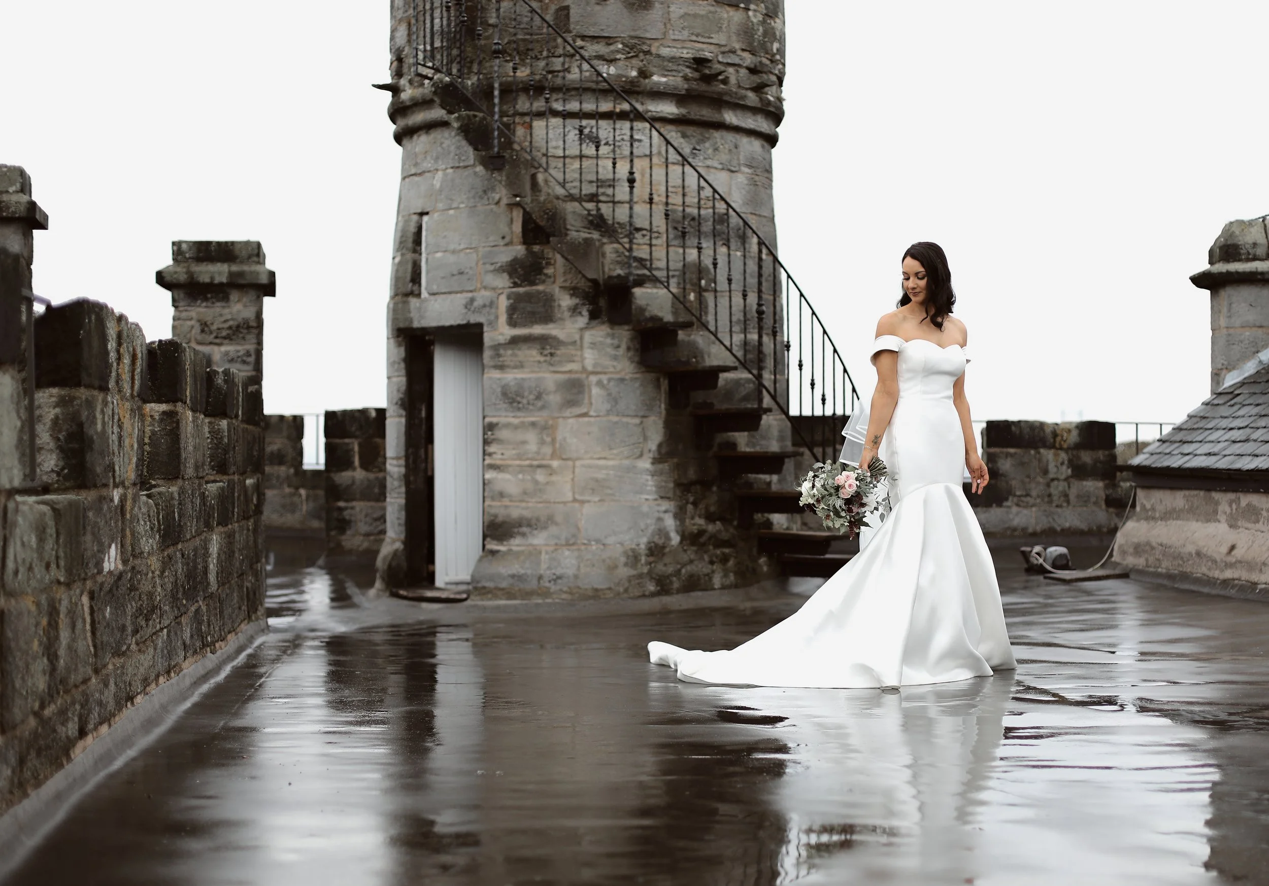 Bride in white gown on wet stone terrace with old stone tower background.