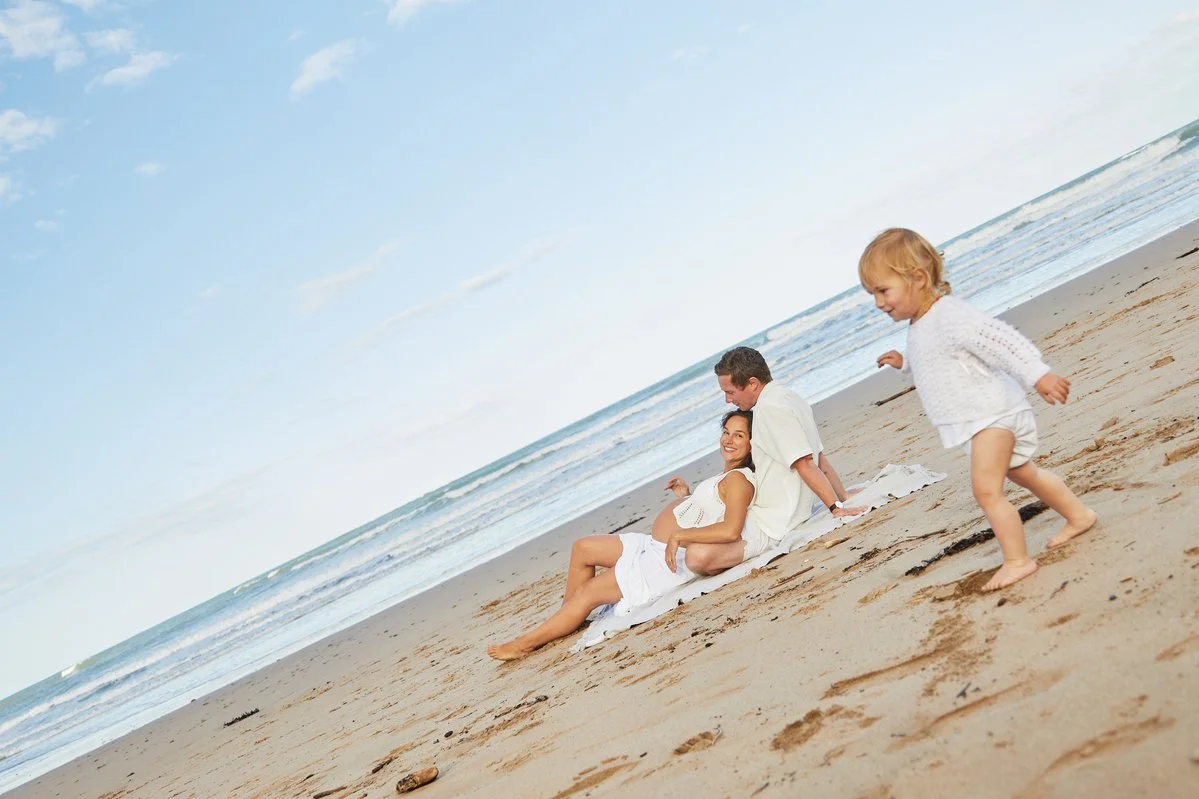 Family relaxing on a beach with a child running, a man and woman sit together in the sand; ocean in the background.