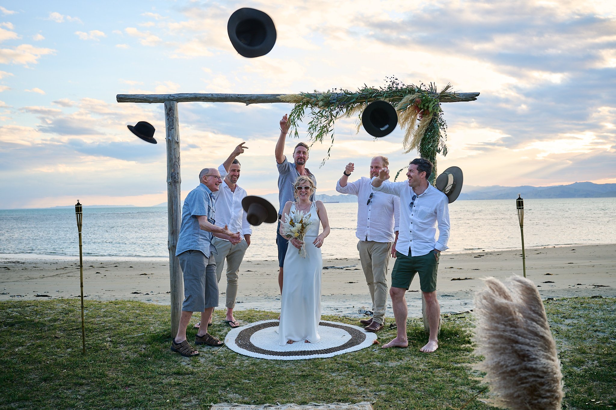 Beach wedding celebration with bride and men tossing hats in the air.