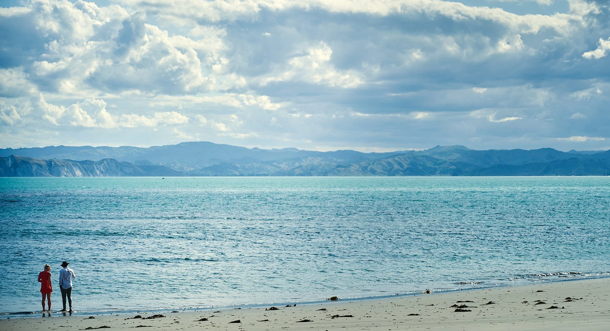 Two people walking along a beach with ocean and mountains in the background under a cloudy sky.