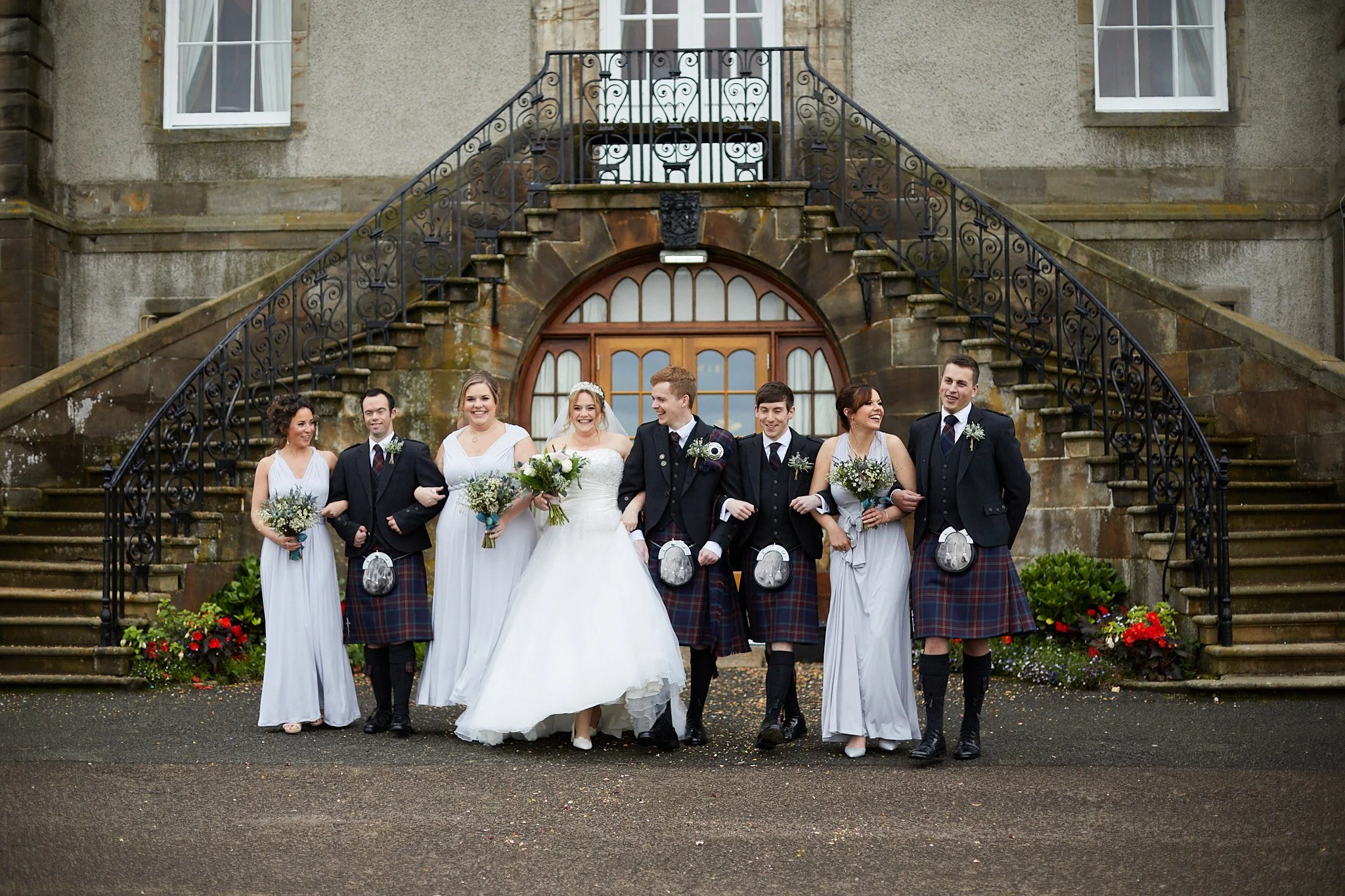 Wedding party with bridal gowns and kilts in front of a stone building.