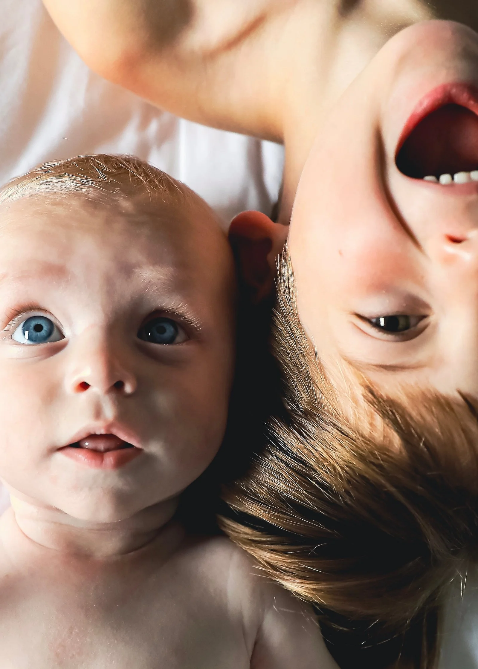 Close-up of a baby lying next to an older child, both with light skin, eyes open, and the older child smiling.