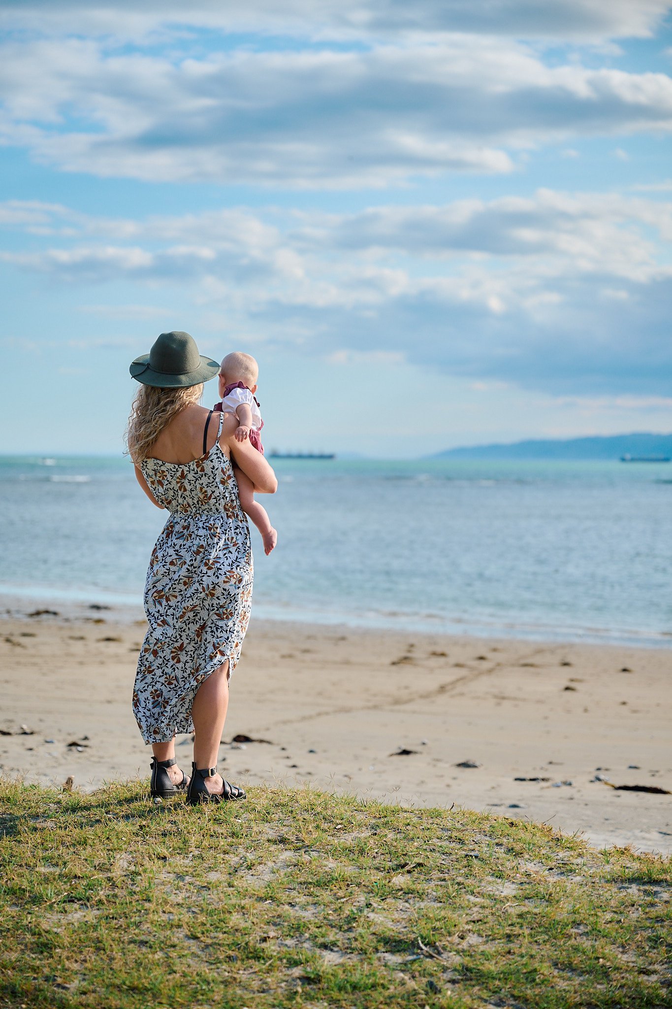 A woman in a floral dress and hat holding a baby, standing on a grassy area by the beach with the ocean in the background.