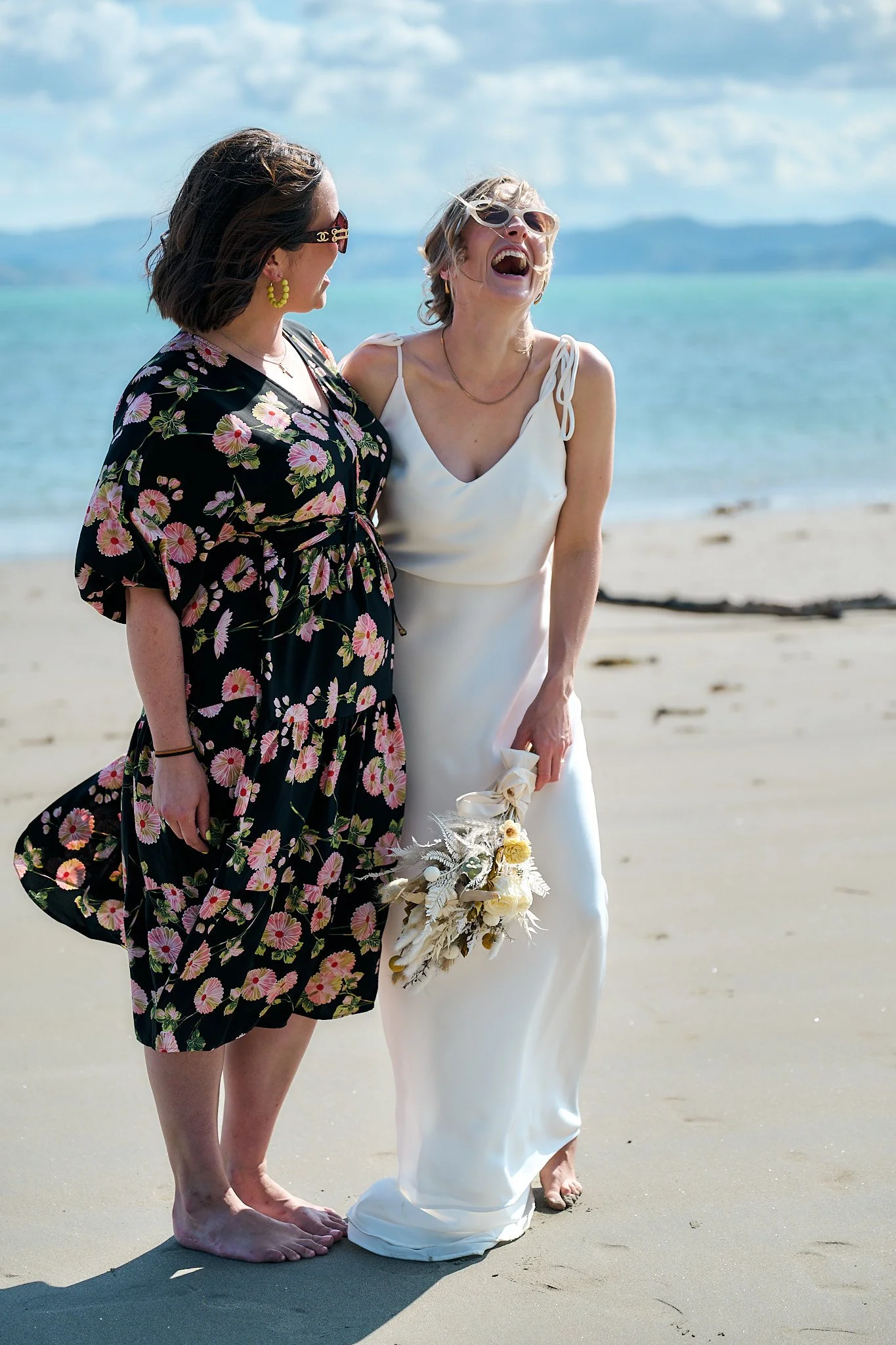 Two women laughing together on a beach; one in a floral dress, the other in a white dress holding a bouquet.