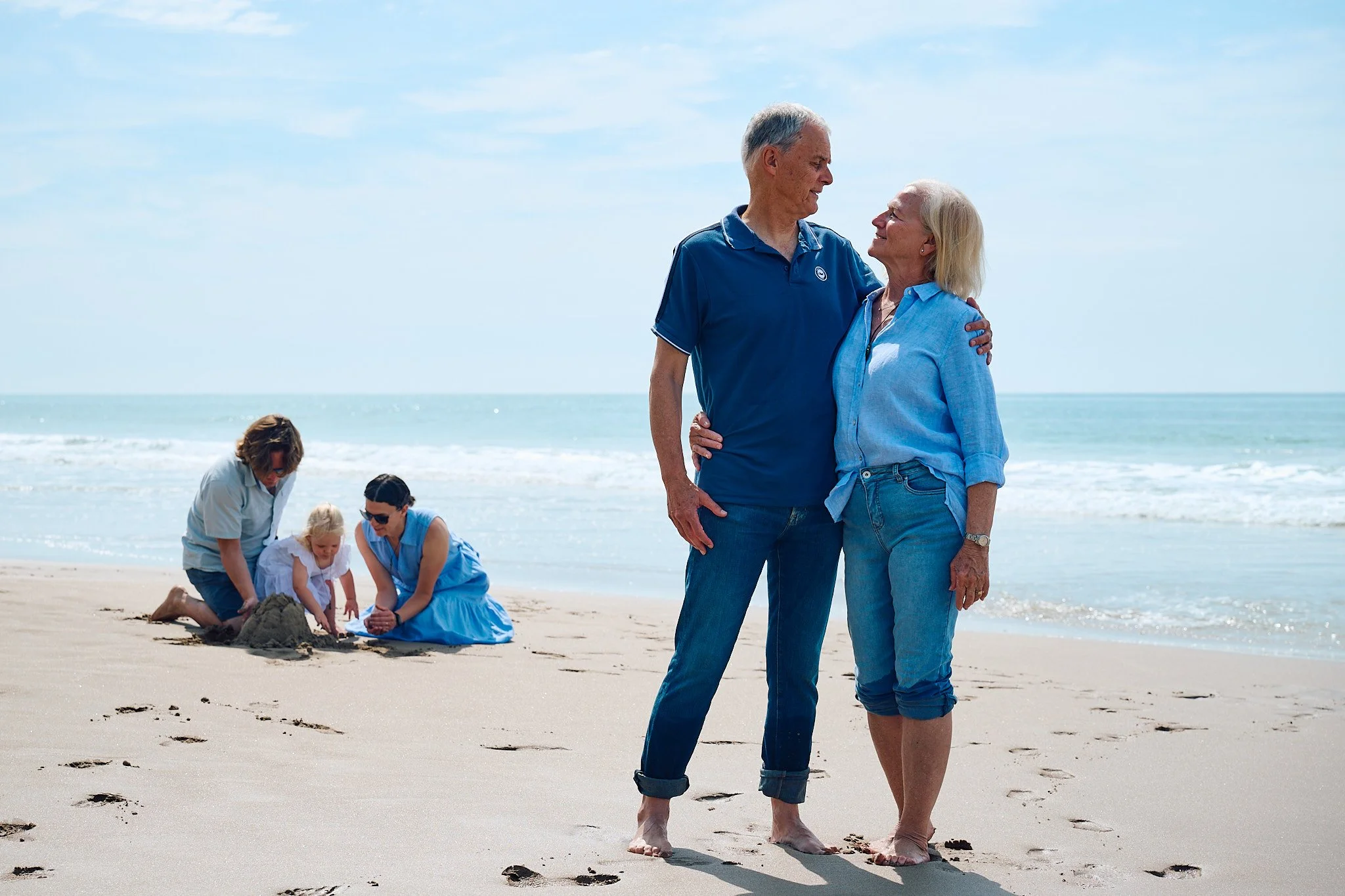 Two couples at the beach, one older couple embracing, and one family with a child building a sandcastle.
