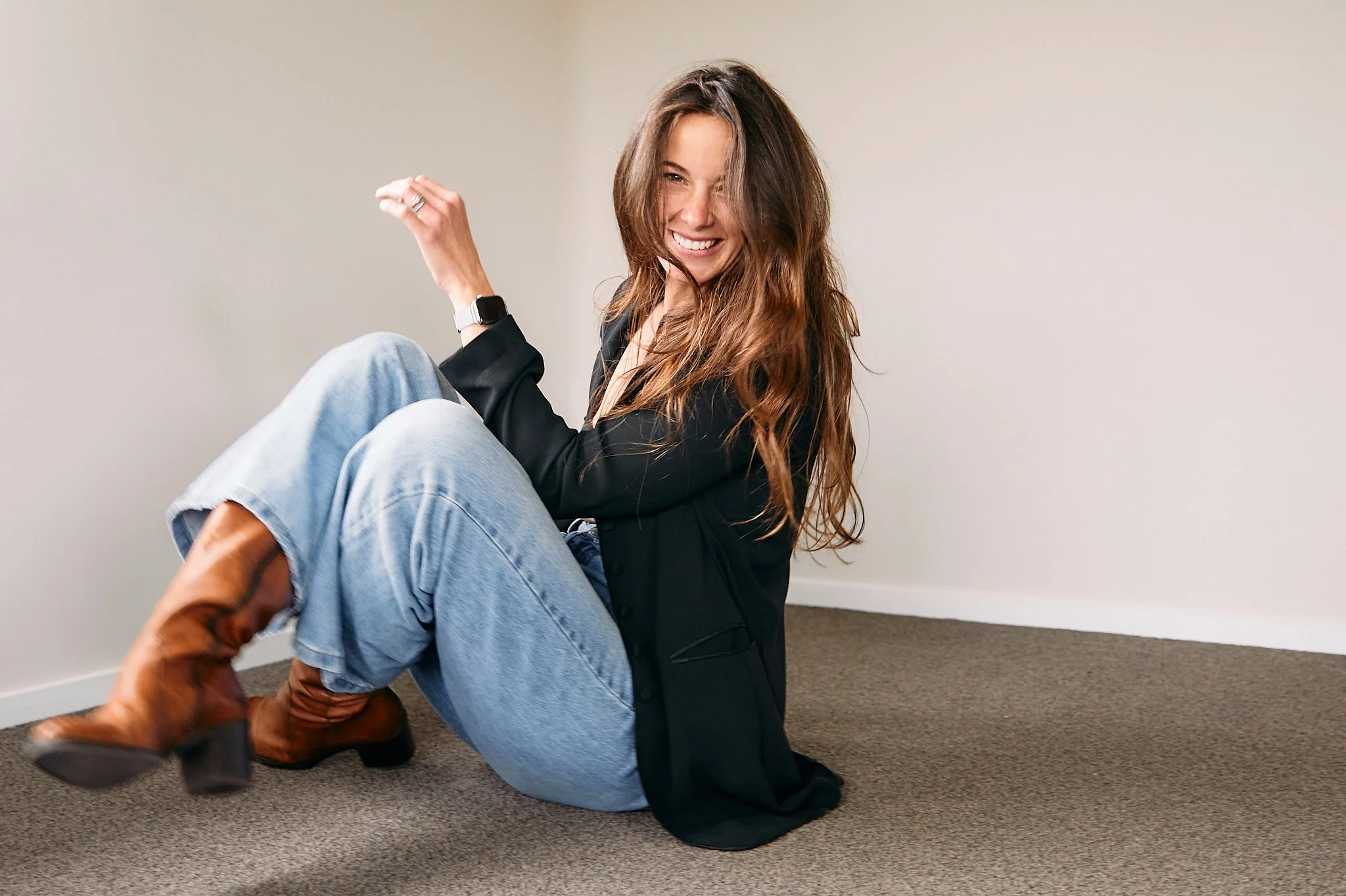 Smiling woman sitting on floor wearing jeans, black jacket, and brown boots.