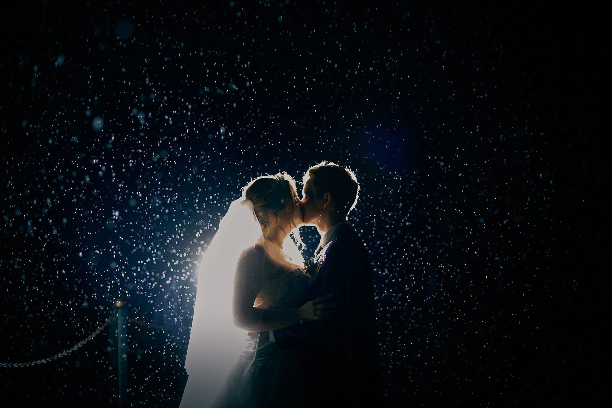 Silhouette of a bride and groom kissing with rain droplets illuminated in the dark background.