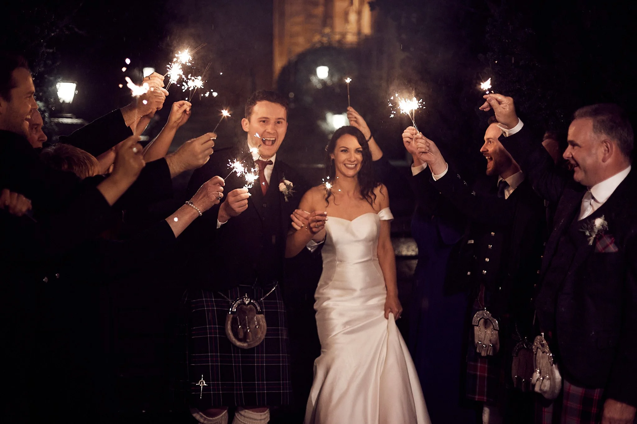 Bride and groom in wedding attire holding sparklers surrounded by guests at night.