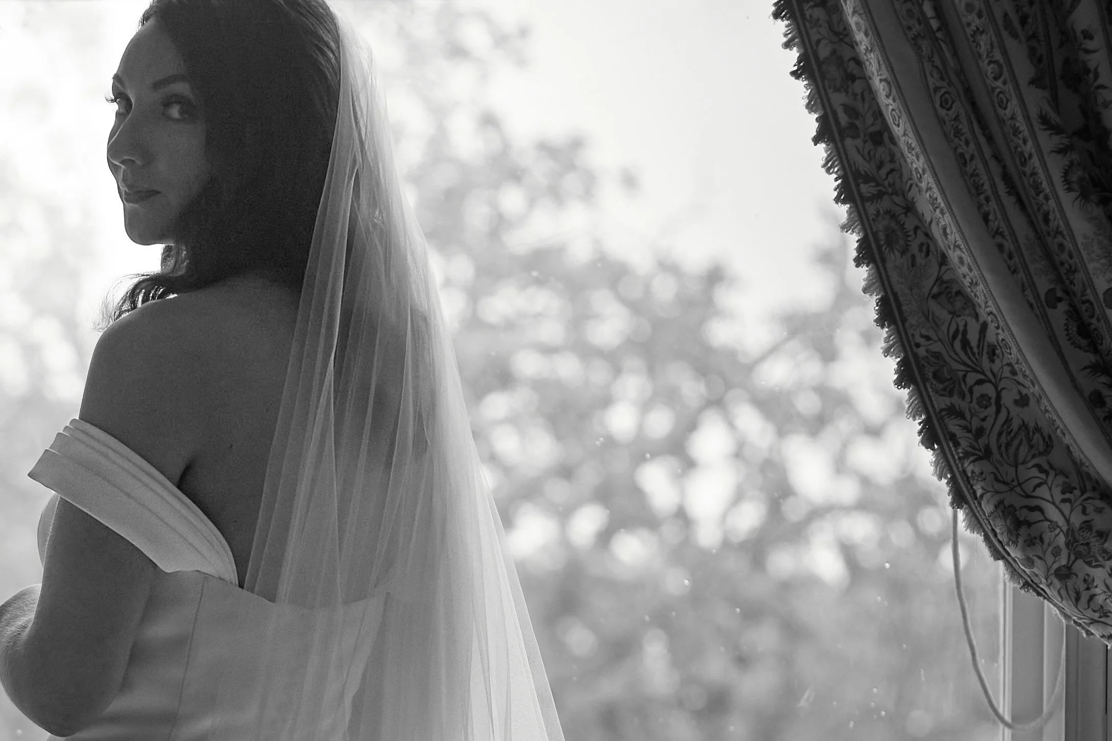 Black and white photo of a bride in a wedding dress with a veil, standing near a window with patterned curtains.