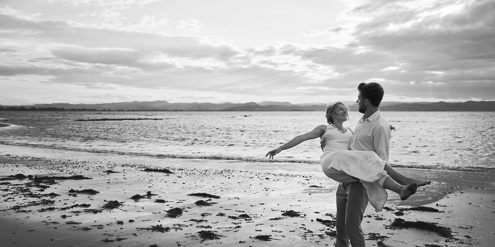 Black and white photo of a man carrying a woman on a beach.
